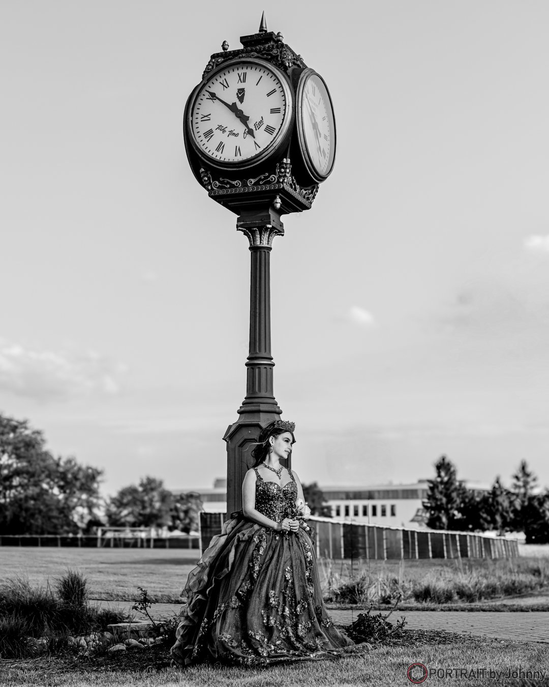 A black-an-white Quinceanera portrait of woman in an ornate gown and tiara standing on grass, holding flowers, beneath a tall clock tower showing the time as 4:52.