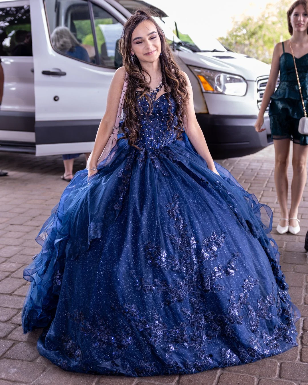 A young woman in a formal, dark blue ball gown with floral embellishments standing on a brick pavement, with a white vehicle and other women in dresses in the background.