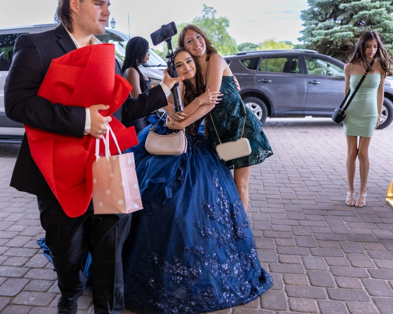 Group of young women dressed in formal gowns, taking a selfie outdoors near cars, with one woman hugging another, while a young man holds gift bags and a large gift.