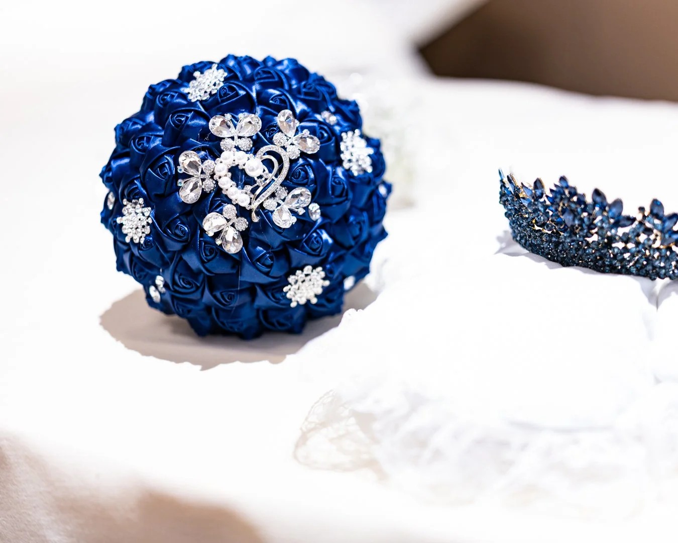 A blue bouquet of artificial roses decorated with rhinestone butterfly and snowflake pins, and a separate blue rhinestone tiara, placed on white fabric.