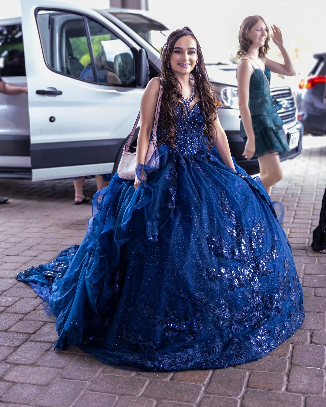 Young woman in a stunning, elaborate blue ball gown with sequins and lace, smiling at a formal event outside, with another woman in a similar dress in the background.