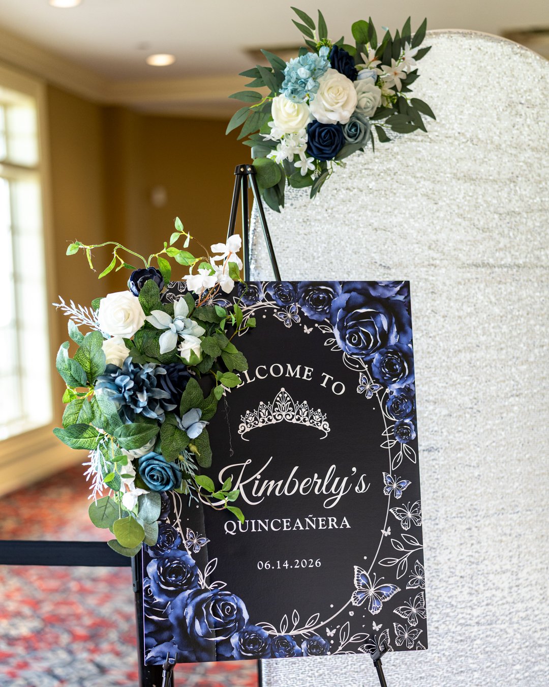 Decorative welcome sign for Kimberly's Quinceañera with blue roses, white flowers, and greenery, displayed on a black easel.