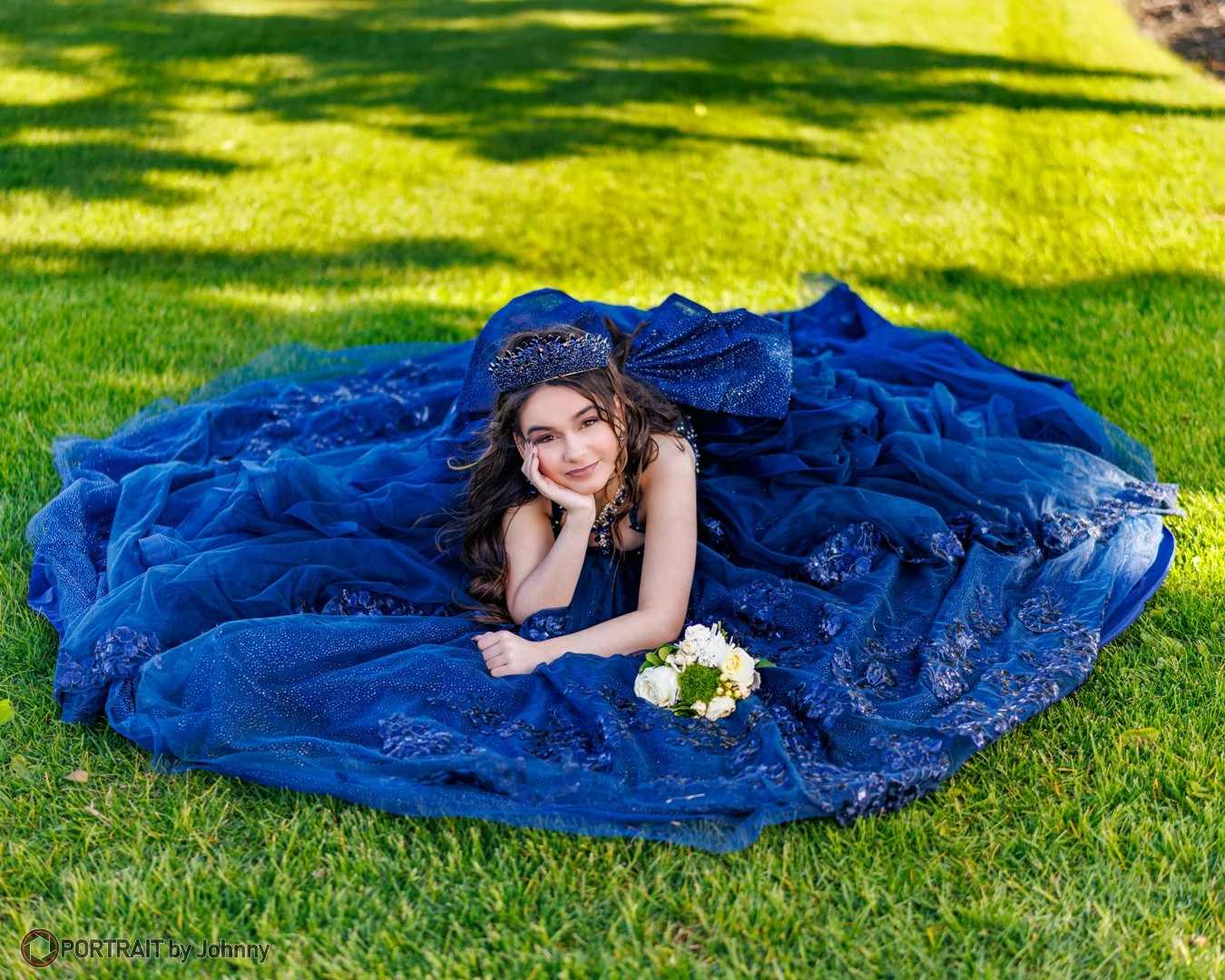 Quinceanera portrait of young woman in a blue gown and tiara lying on grass with a bouquet of white flowers.