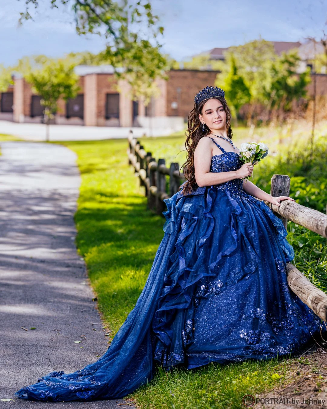 Quinceanera portrait of young woman in a royal blue formal gown holding a bouquet of white roses, standing outdoors near a wooden fence with trees and a building in the background.
