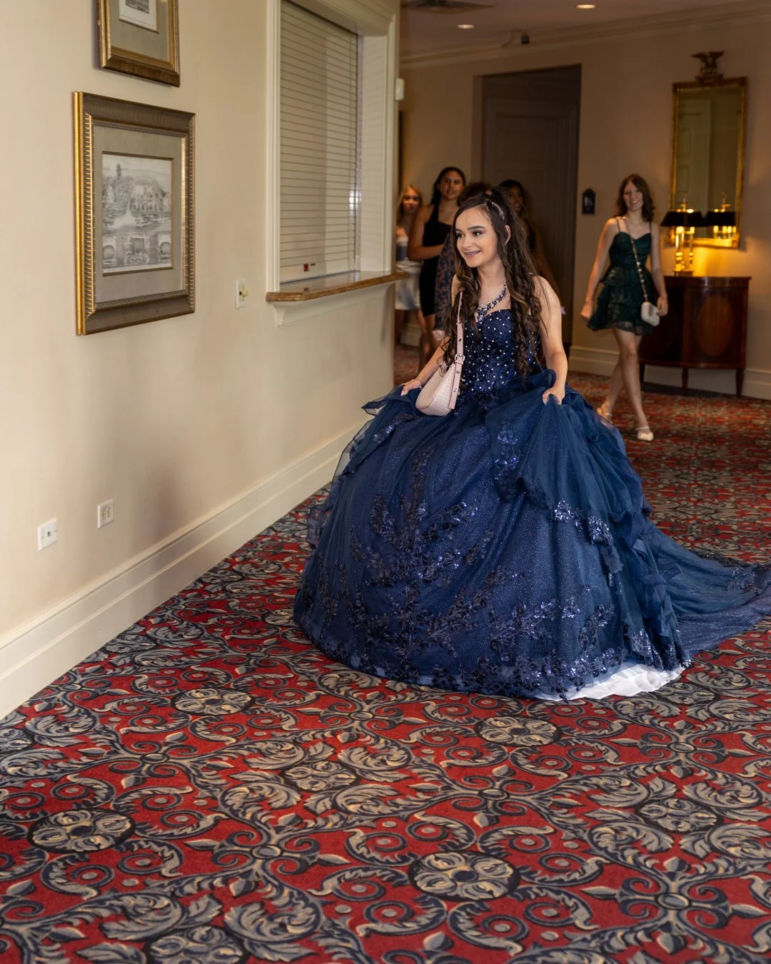Young woman in a dark blue formal gown sitting on the floor in a hallway, with a group of girls in the background.
