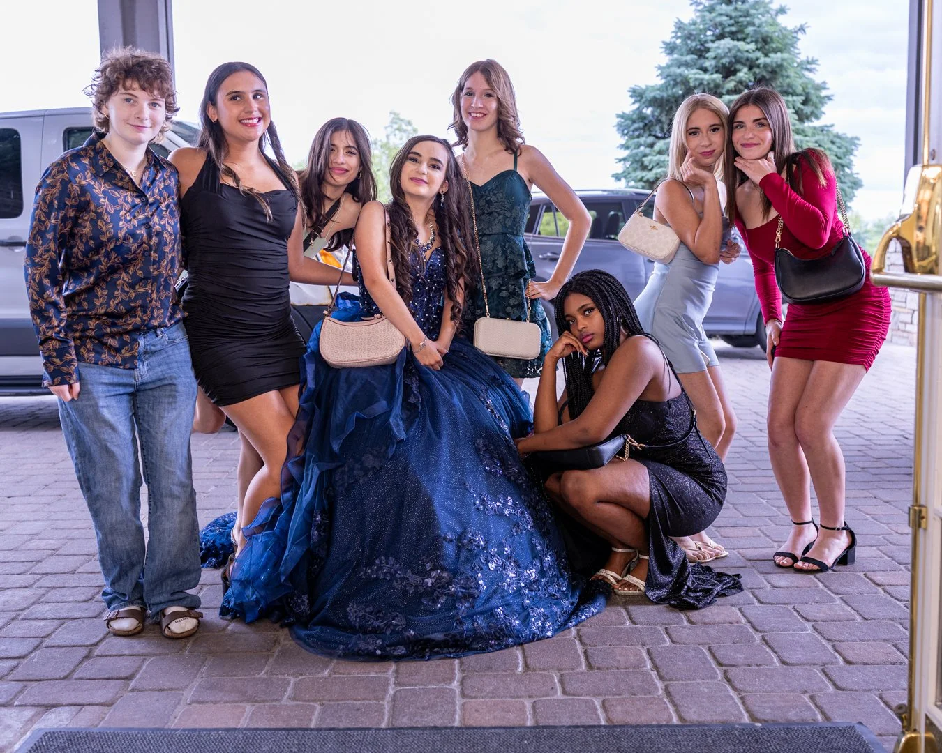 Group of nine young women dressed in formal and casual outfits posing outside near cars, with one woman in an elaborate blue gown sitting on the ground.