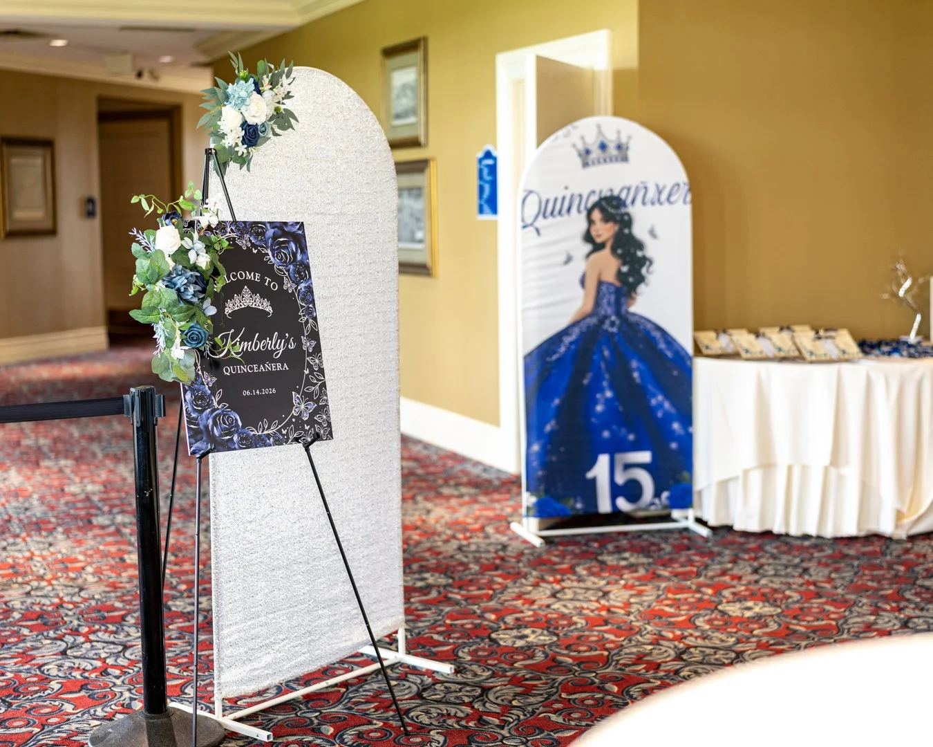 Decorative sign with flowers and butterfly welcoming guests to Kimberly's quinceañera on June 14, 2026, next to a large illustrated poster of a girl in a blue dress celebrating her 15th birthday.