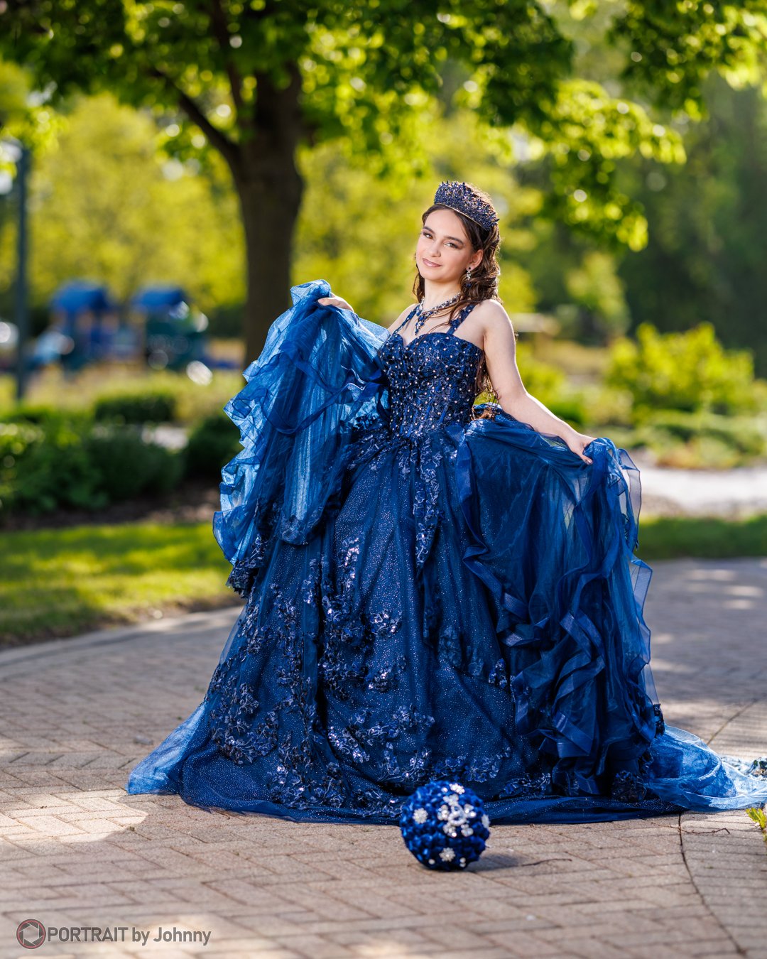 Quinceanera portrait of young woman in a flowing blue gown and tiara standing outdoors on a brick path, holding up her dress with one hand, with a bouquet on the ground in front of her, under a large tree, with sunlight filtering through the leaves.