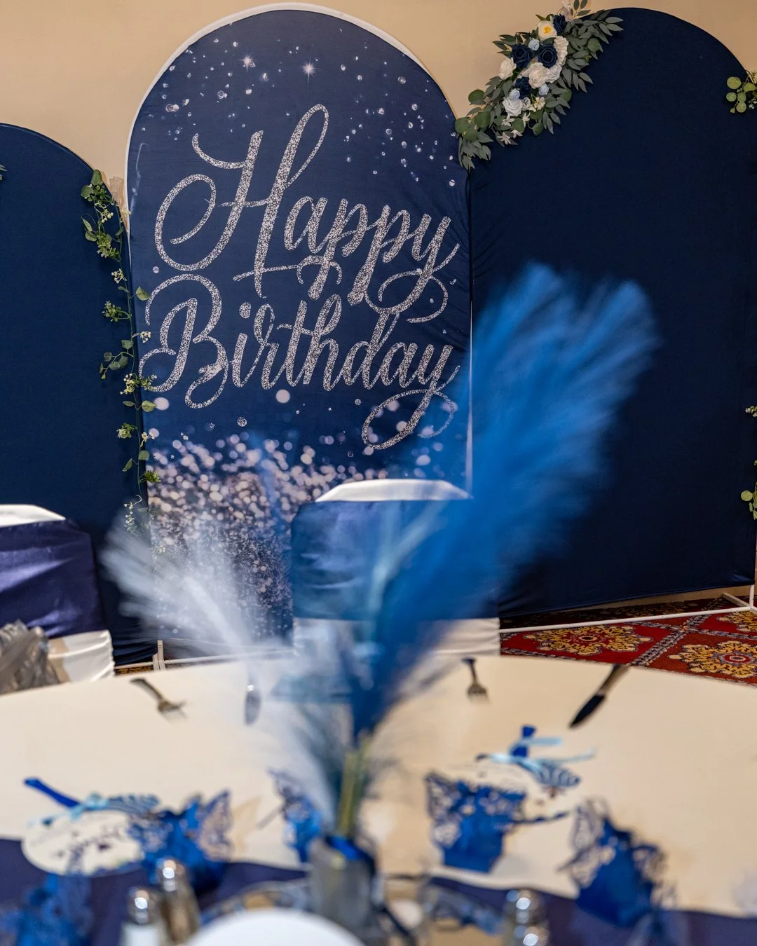 Decorative backdrop with 'Happy Birthday' written in script, surrounded by blue, white, and green floral accents, set up for a celebration.