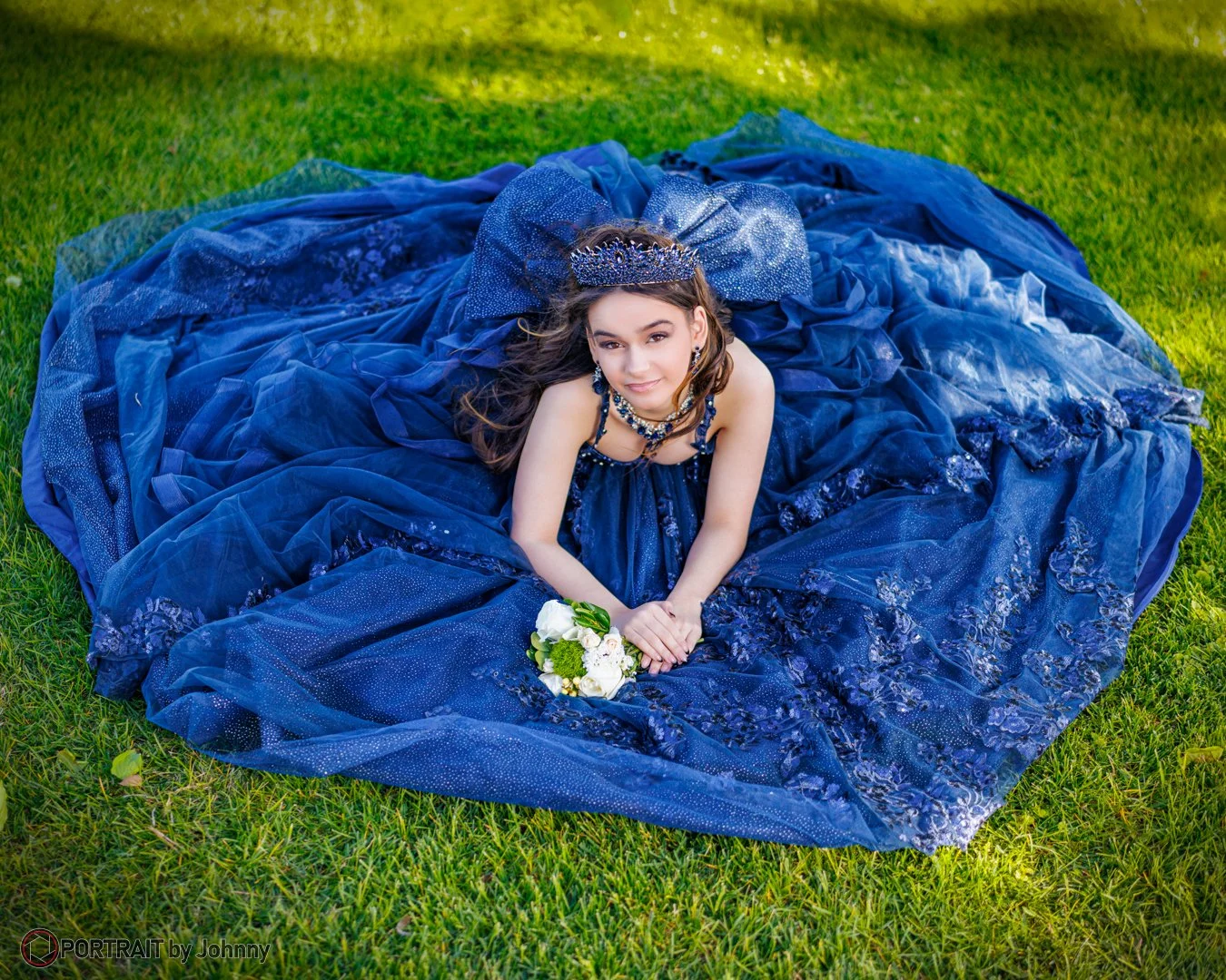 Quinceanera portrait of young girl in a royal blue ball gown, wearing a tiara and jewelry, lying on grass with a bouquet of white flowers.