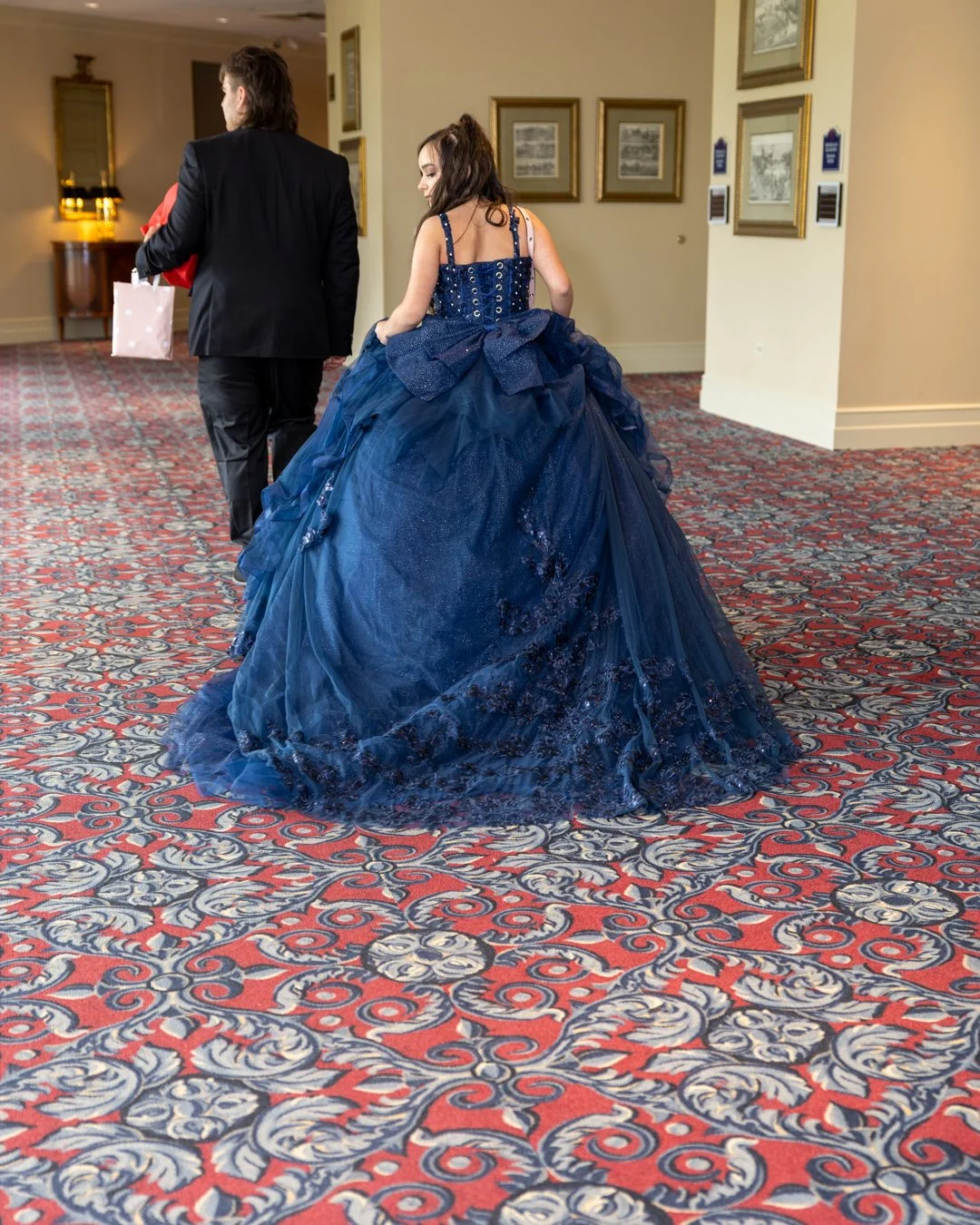 A woman in a dark blue ball gown walking on a patterned carpet, with a man in a black suit walking ahead of her.