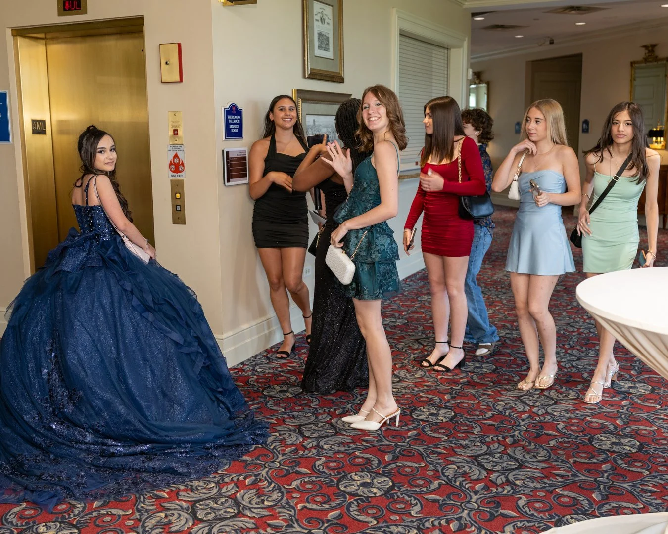 Young women dressed in formal dresses line up to take photos with a girl in a large, blue ball gown at an indoor event.