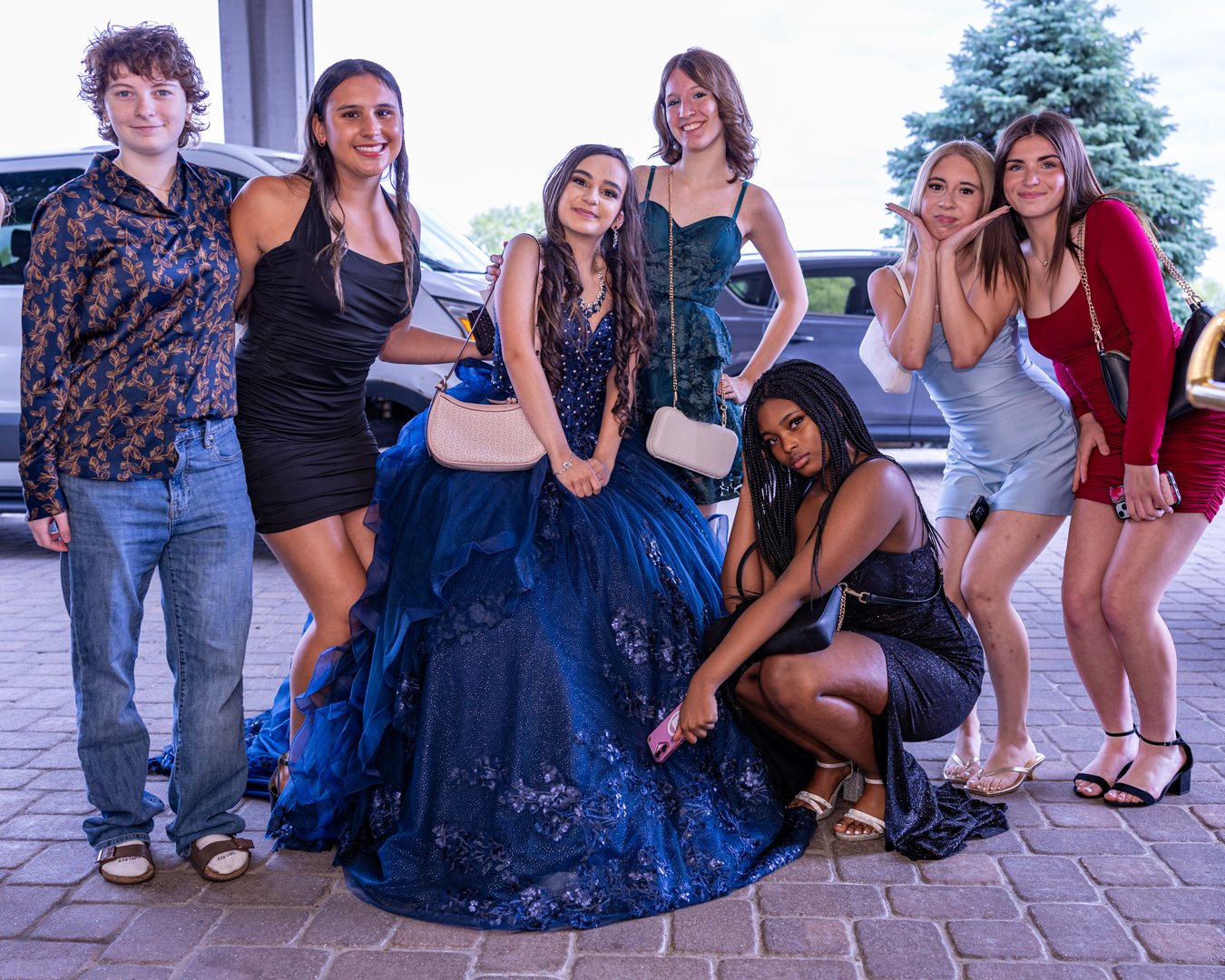 Seven young women pose together outdoors for a group photo, with cars and trees in the background. One woman is seated in a dark blue fancy gown, while the others are standing or kneeling around her in stylish dresses and casual wear.