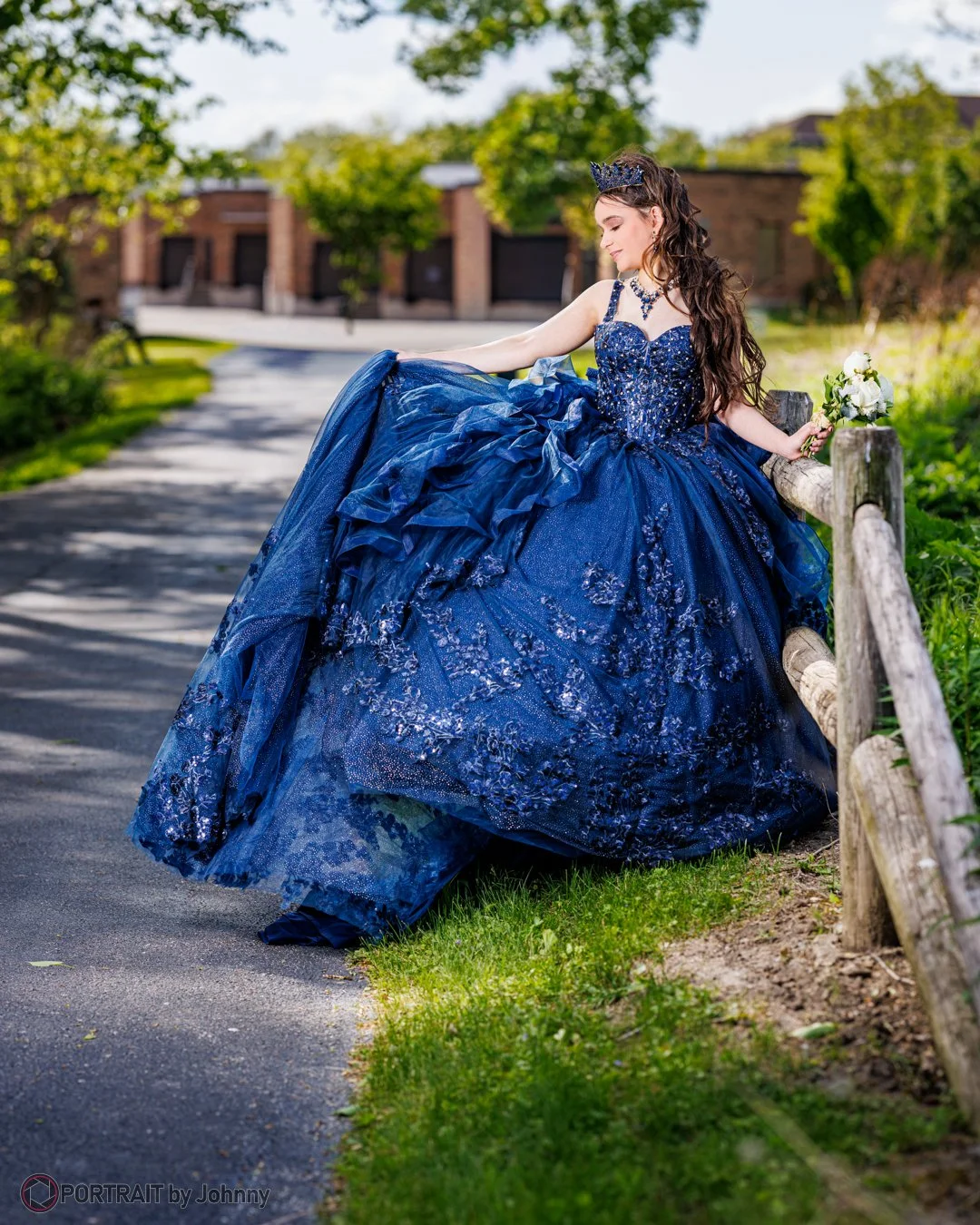 Quinceanera portrait of young woman in a royal blue, intricately embroidered ball gown and matching crown, sitting on a wooden fence along a park path, holding a small bouquet of white flowers.