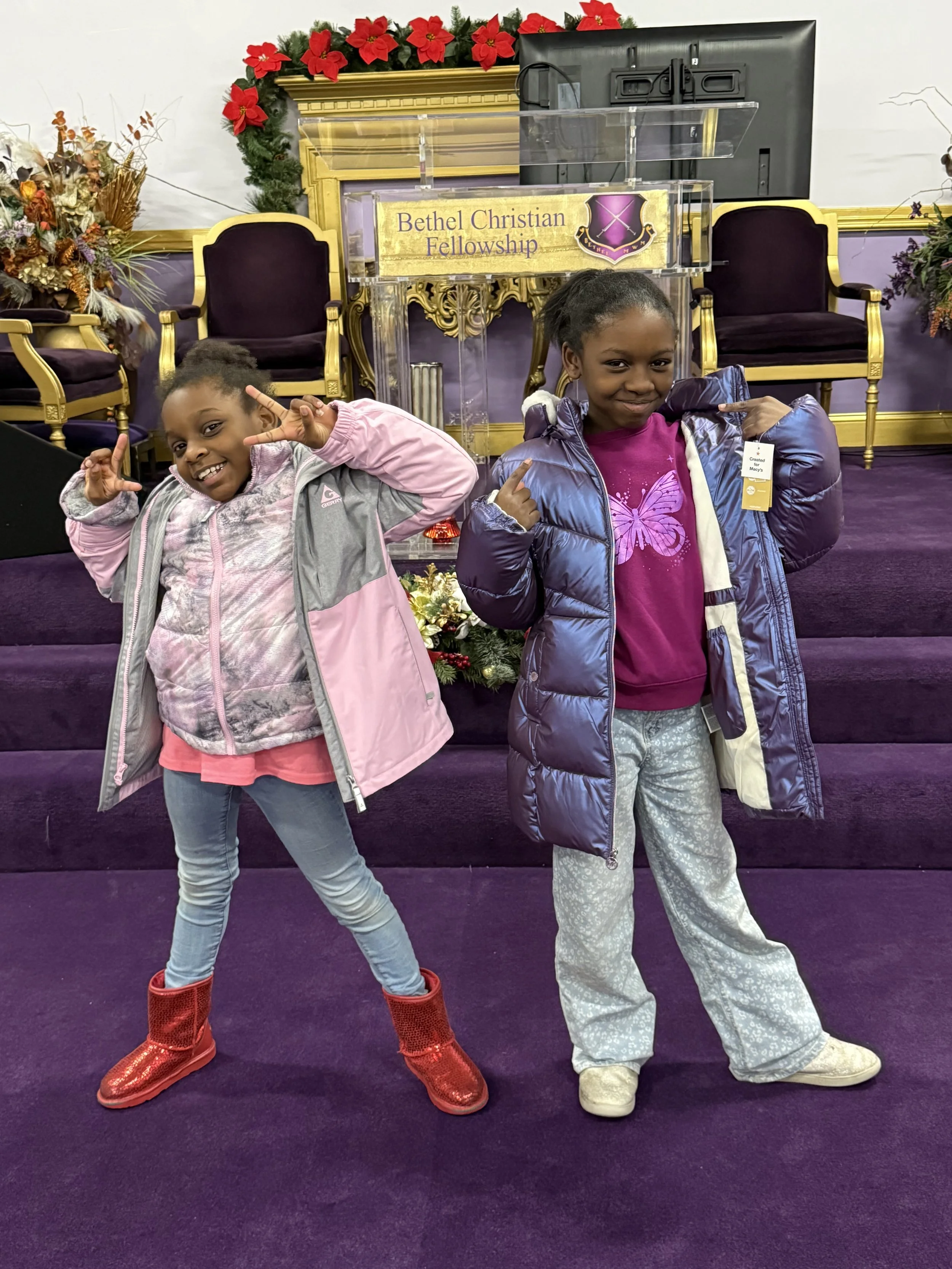 Two smiling young girls posing on a purple carpeted stage with church chairs and a lectern labeled Bethel Christian Fellowship in the background. They are wearing colorful jackets and boots, striking playful poses.
