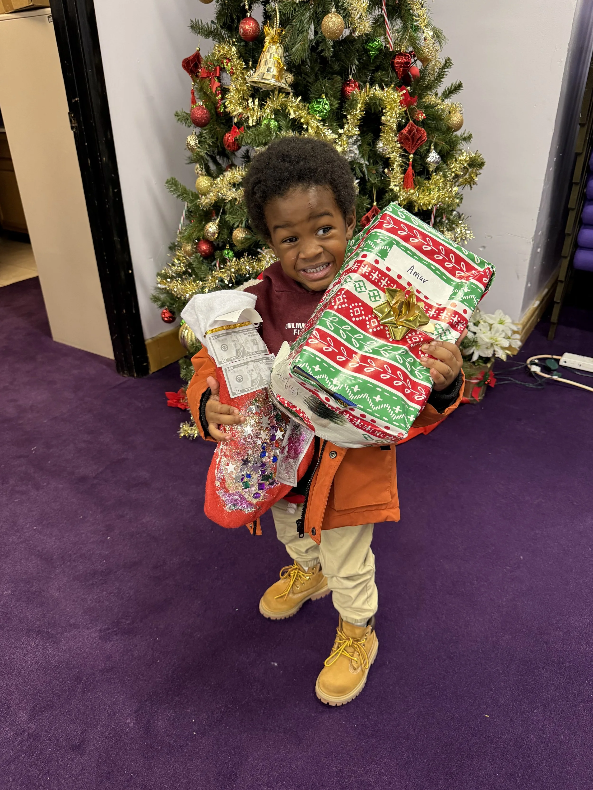 A young boy stands in front of a decorated Christmas tree, holding a wrapped gift and a Christmas stocking, with a joyful expression.