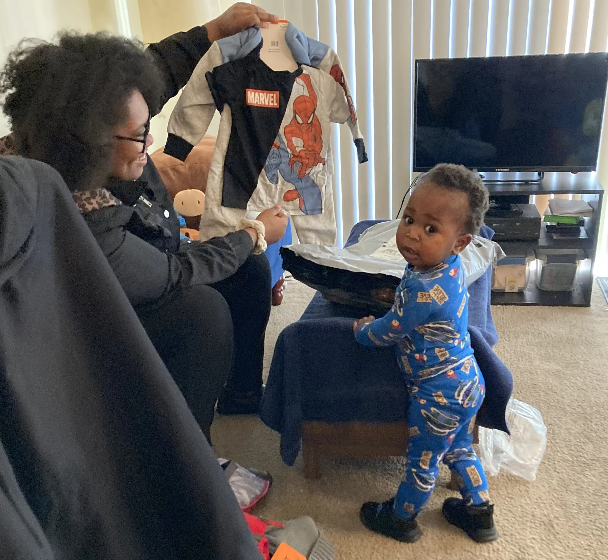 A woman and a young child in a living room. The woman is sitting on a chair, smiling, while another person holds up a Spider-Man themed hoodie. The child is standing on the floor, wearing blue pajamas with cartoon characters, looking at the camera with a curious expression.