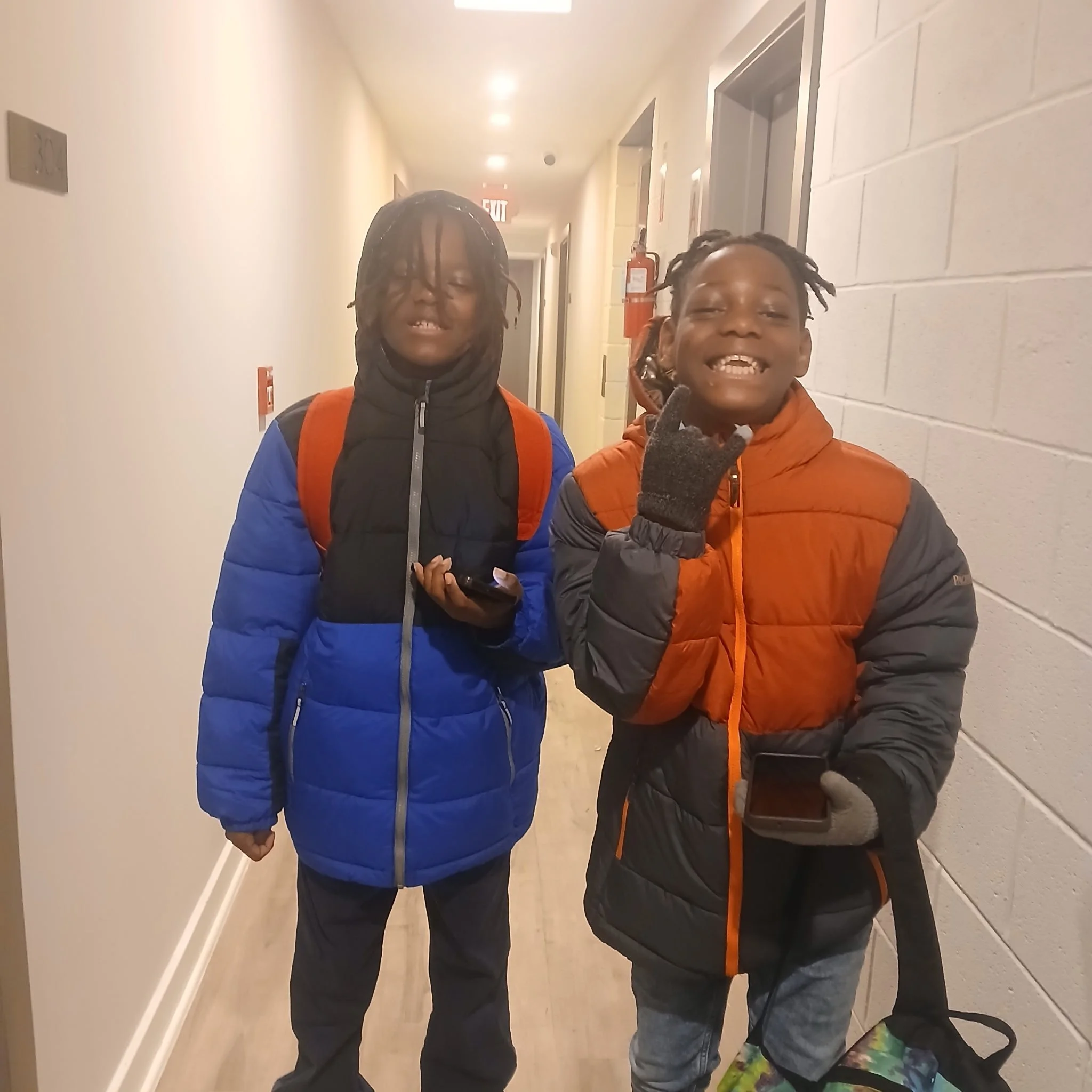 Two young boys standing in a hallway, smiling and holding smartphones. One boy is wearing a colorful puffer jacket and gray gloves, while the other boy is wearing a blue and black jacket with a red backpack.