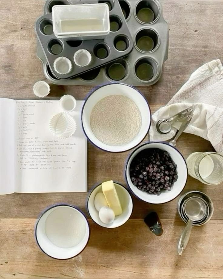 Overhead view of baking ingredients including flour, blueberries, butter, and an egg; muffin tins, measuring cups, paper liners, a glass of water, and an open recipe book on a wooden surface.