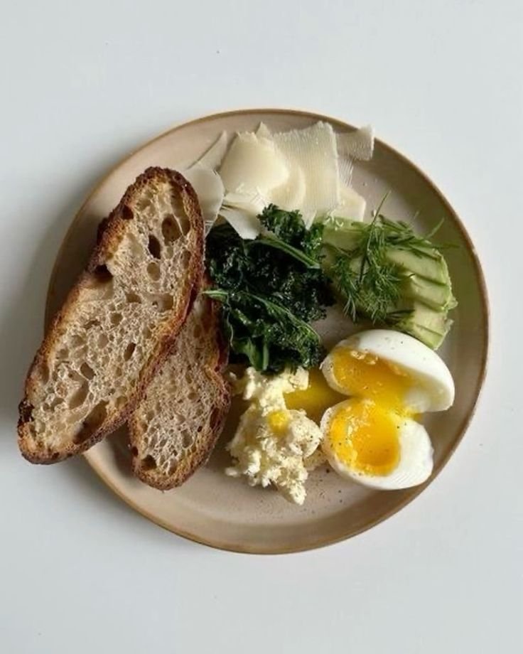 Plate with sliced bread, greens, avocado, shaved cheese, soft-boiled egg, and ricotta cheese.