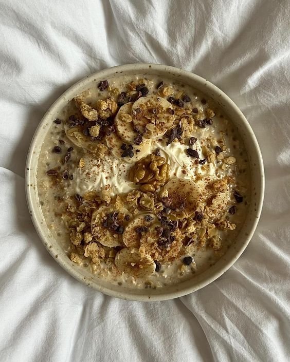 Bowl of oatmeal with banana slices, walnuts, granola, and cocoa nibs, on white bedding.