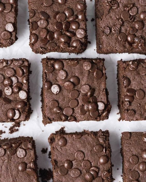 Squares of chocolate brownies with chocolate chips on top, arranged on a white surface.