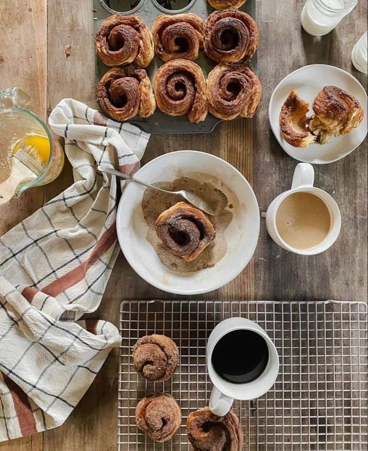 Cinnamon rolls on a cooling rack and in a muffin tin, with coffee and milk on a wooden table.