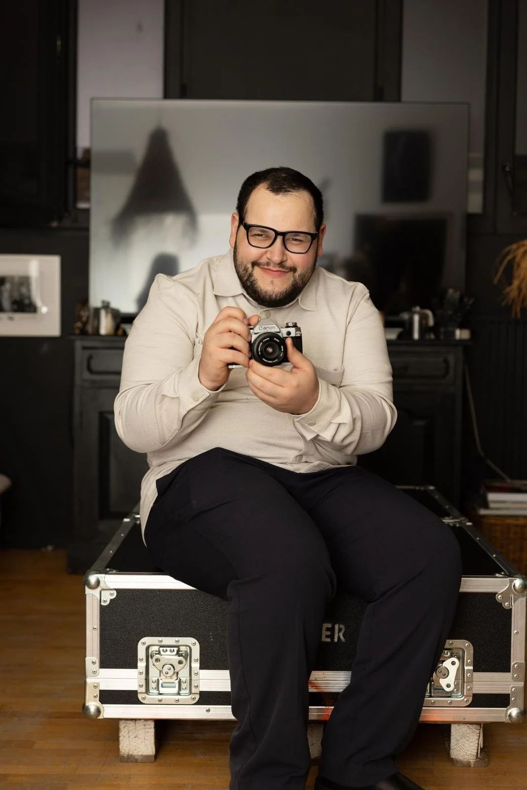 Un homme souriant assis sur une caisse en bois, tenant un appareil photo Canon, dans une pièce avec un grand miroir en arrière-plan.