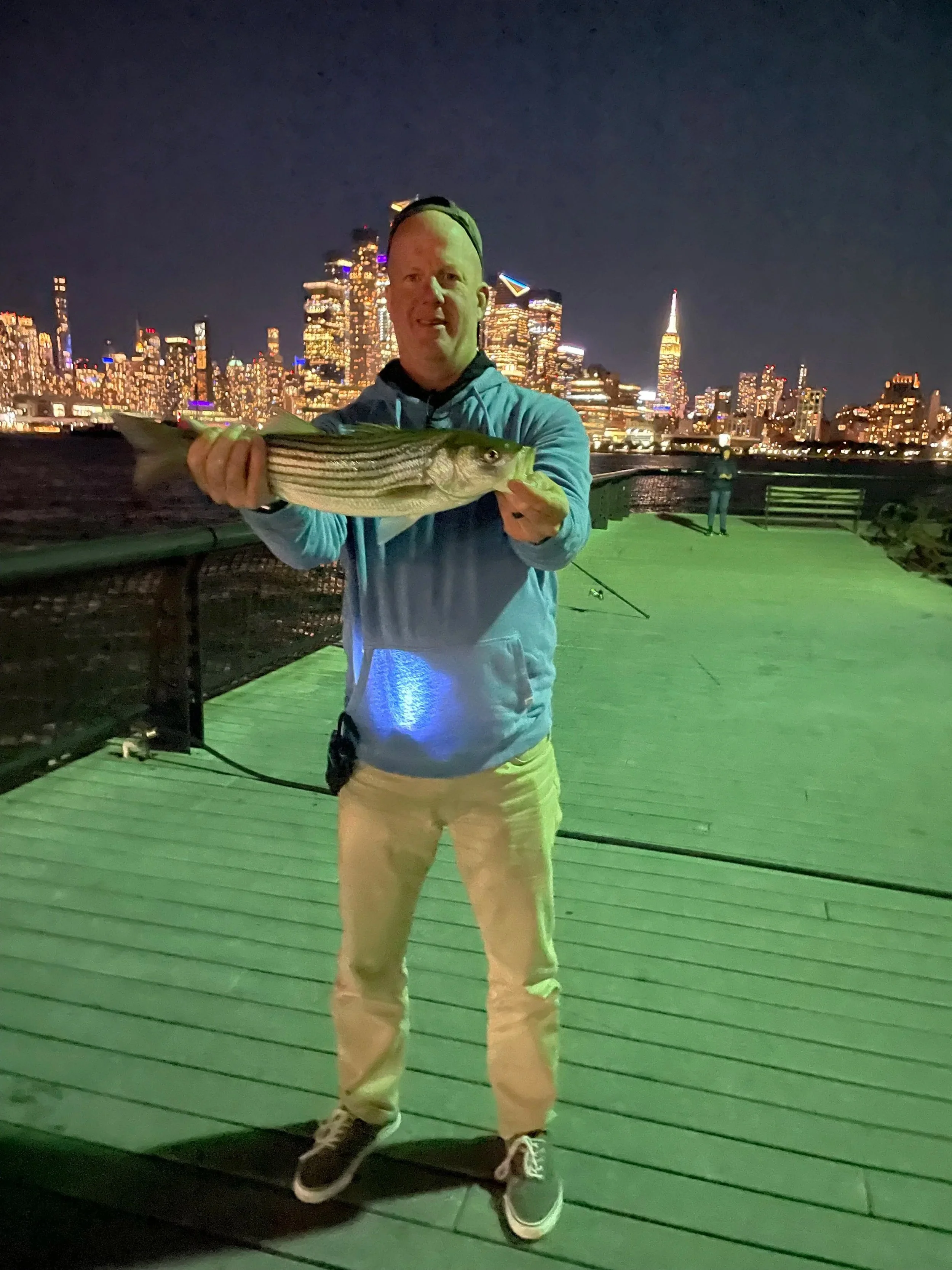 man catching striped bass at night in front of nyc skyline