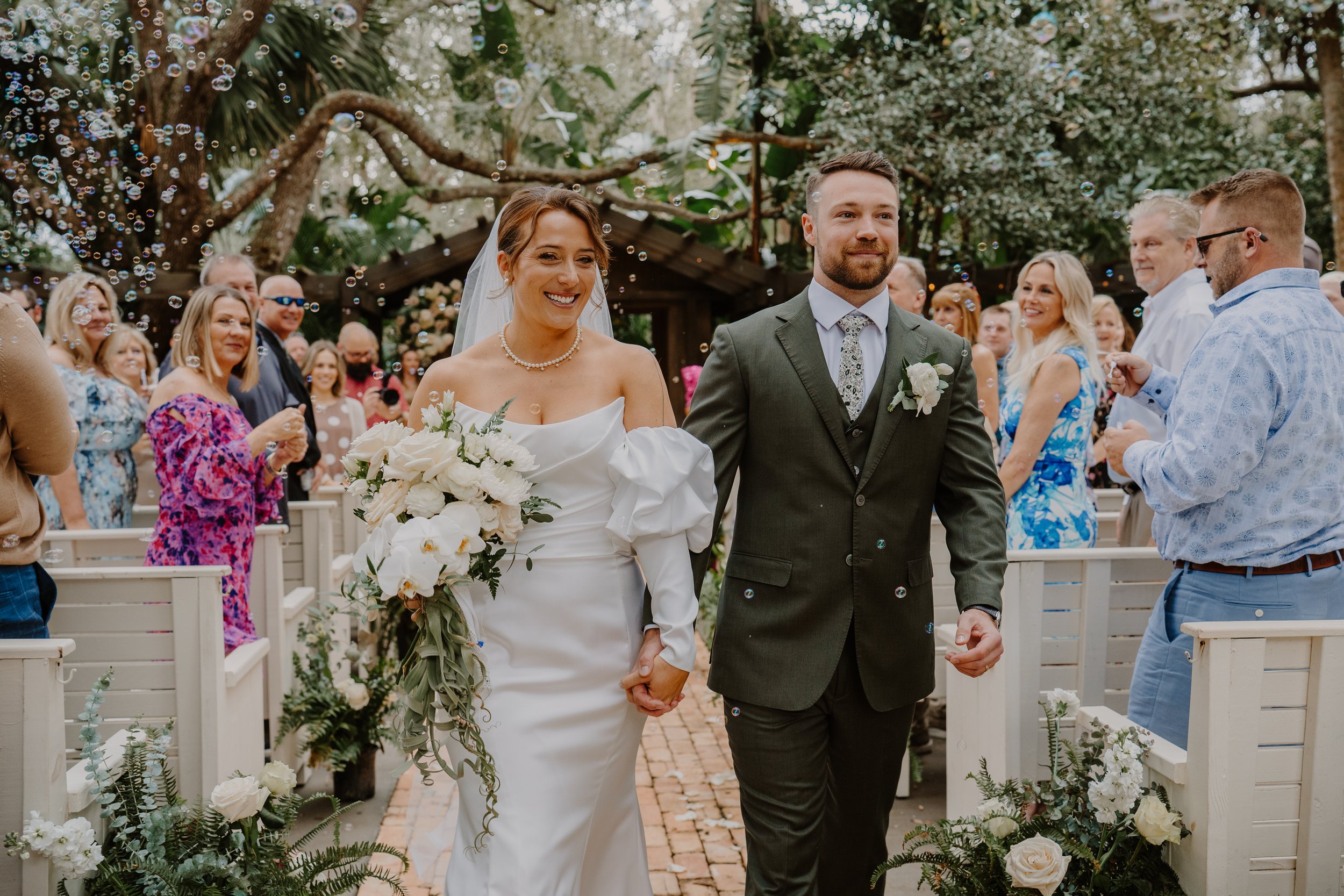 Bride and groom walking down an outdoor aisle, surrounded by guests blowing bubbles, with greenery and flowers in the background.