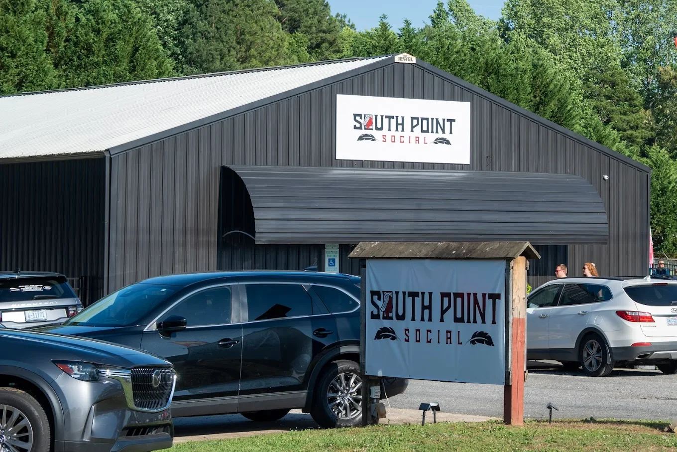 The exterior of a black building with a sign that reads 'South Point Social', with several parked cars and a small signpost in front of the building.