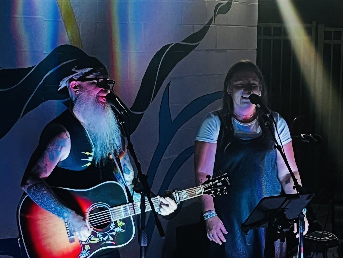 A man with a gray beard, glasses, and tattoos playing an acoustic guitar and singing into a microphone, next to a woman with glasses singing into a microphone, in front of a colorful mural background.