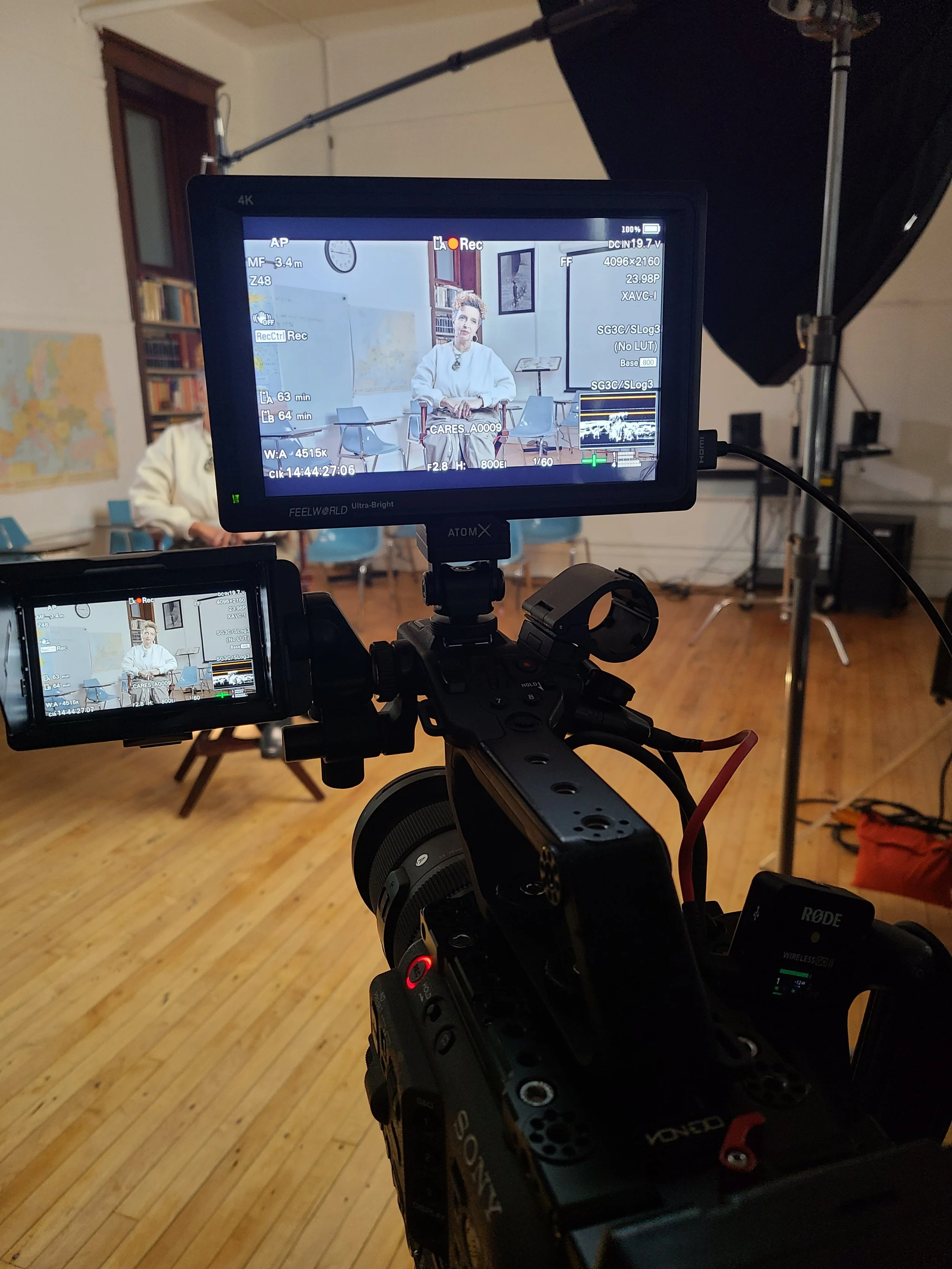 A professional video camera captures a woman seated in front of a classroom setting with chairs, bookshelves, a calendar, and framed pictures on the wall.