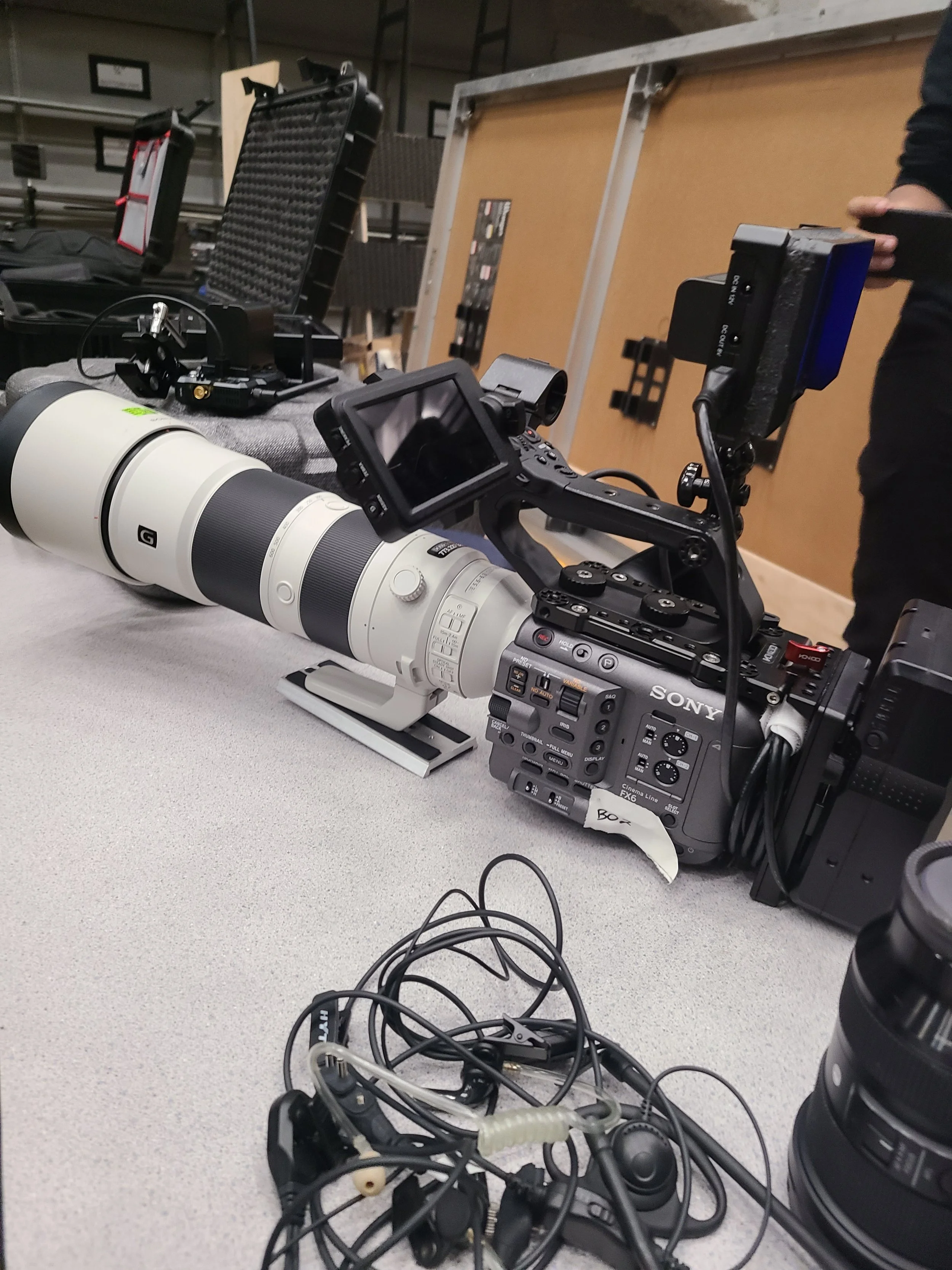 A professional video camera with a large telephoto lens, mounted on a tripod, placed on a grey table, with various electronic accessories and cables in the foreground, and equipment cases and storage racks in the background.