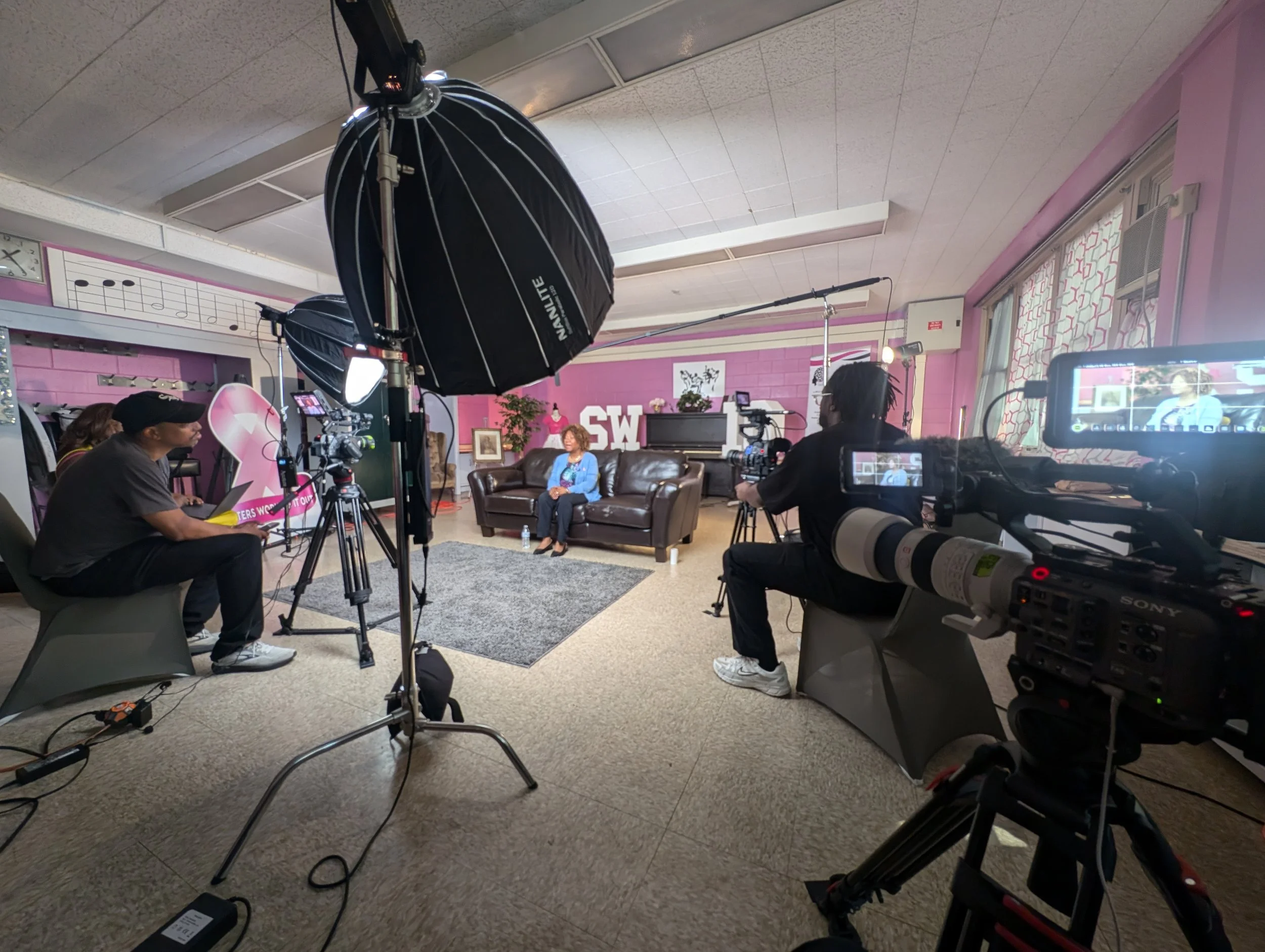 A woman sitting on a brown couch in a studio set, with film crew and cameras around her, pink walls, decorative art, and a large pink and white banner in the background.
