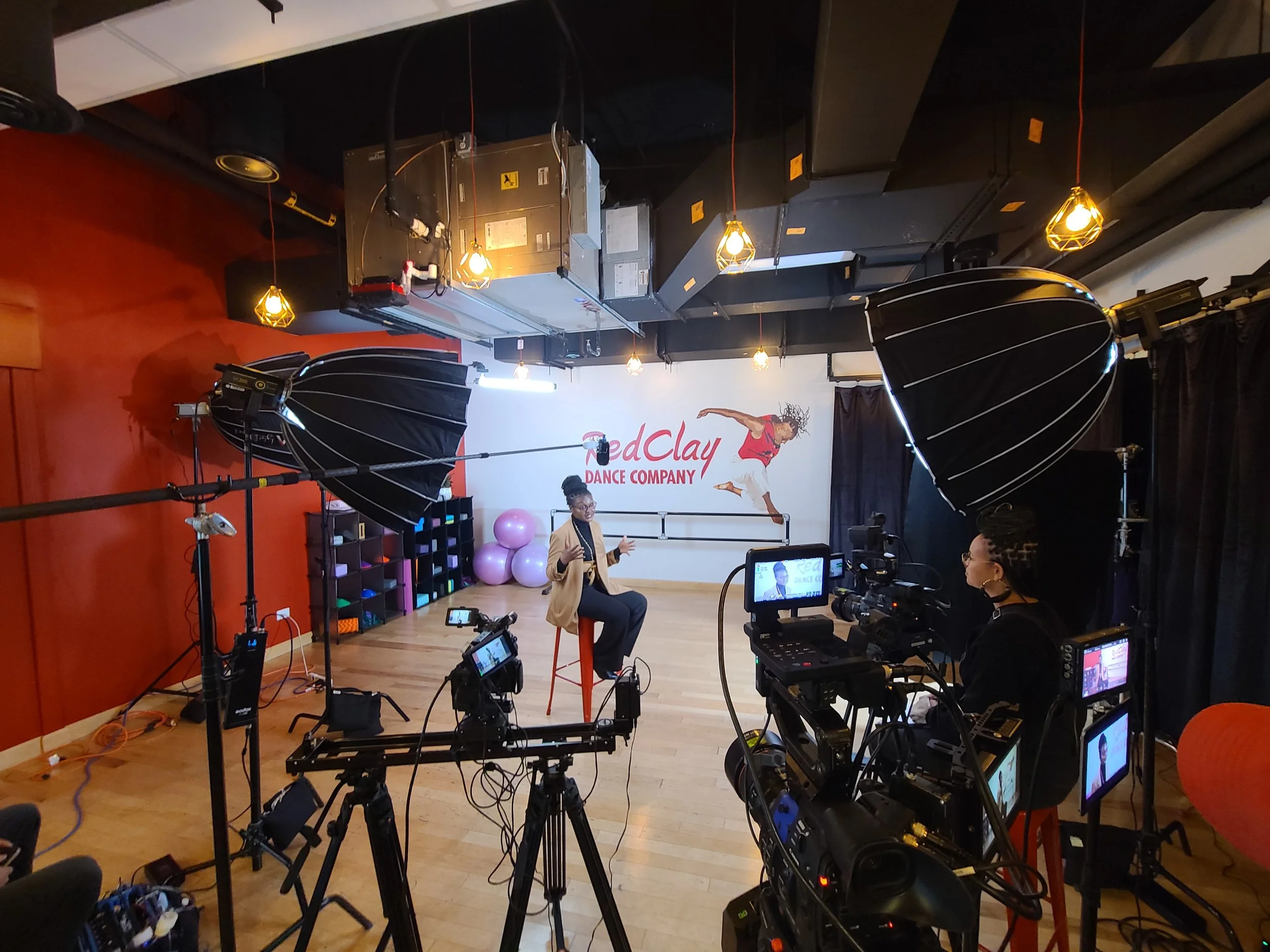 A dance studio with cameras and lighting equipment set up for filming or recording. A woman is sitting on a stool, speaking or giving an interview, with a sign behind her that reads 'RedClay DANCE COMPANY.' The studio has a wooden floor, a white wall
