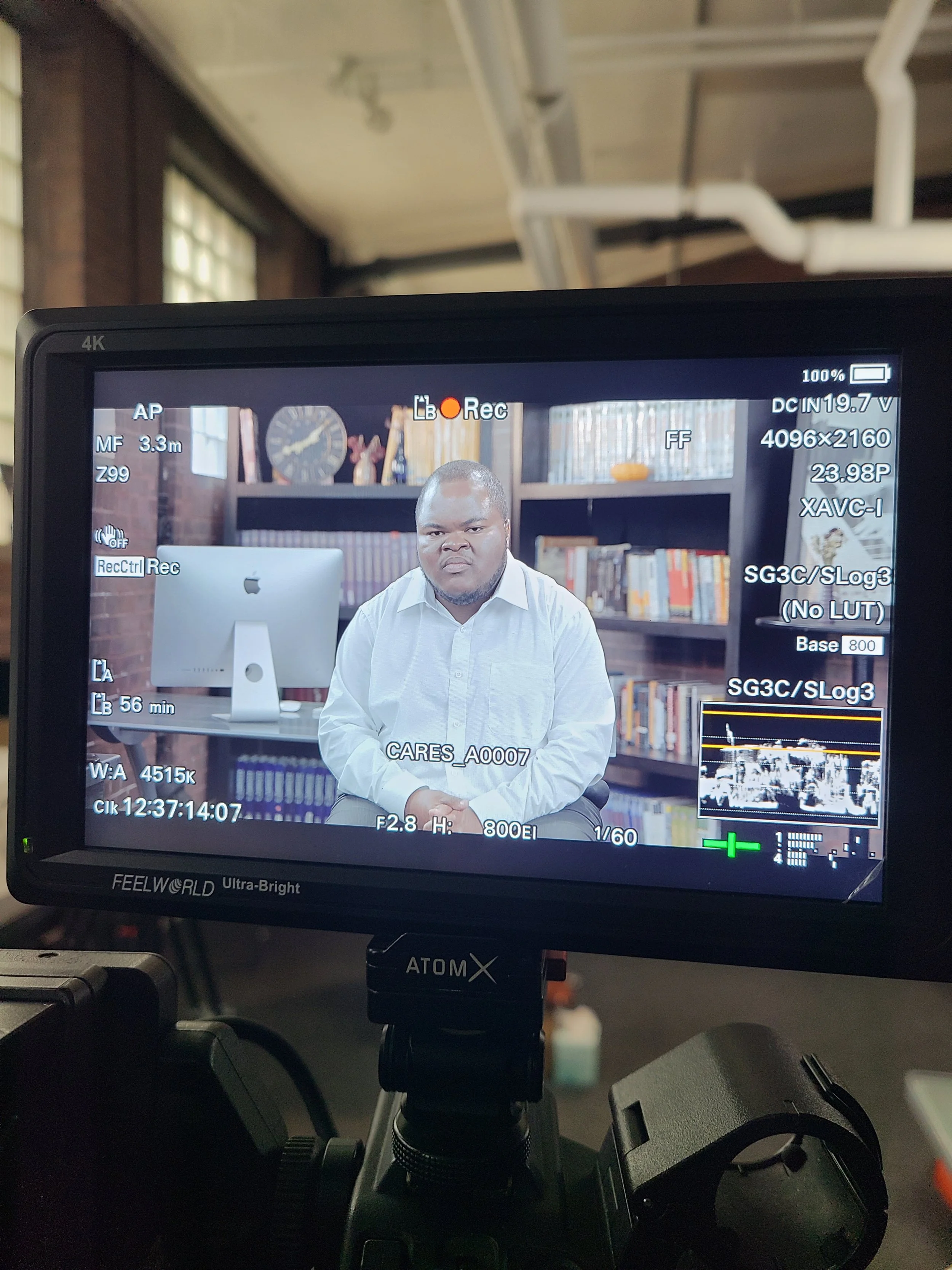 A camera monitor displays a shot of a man in a white shirt sitting in front of a bookshelf with books and decorative items in a room with brick walls and large windows.