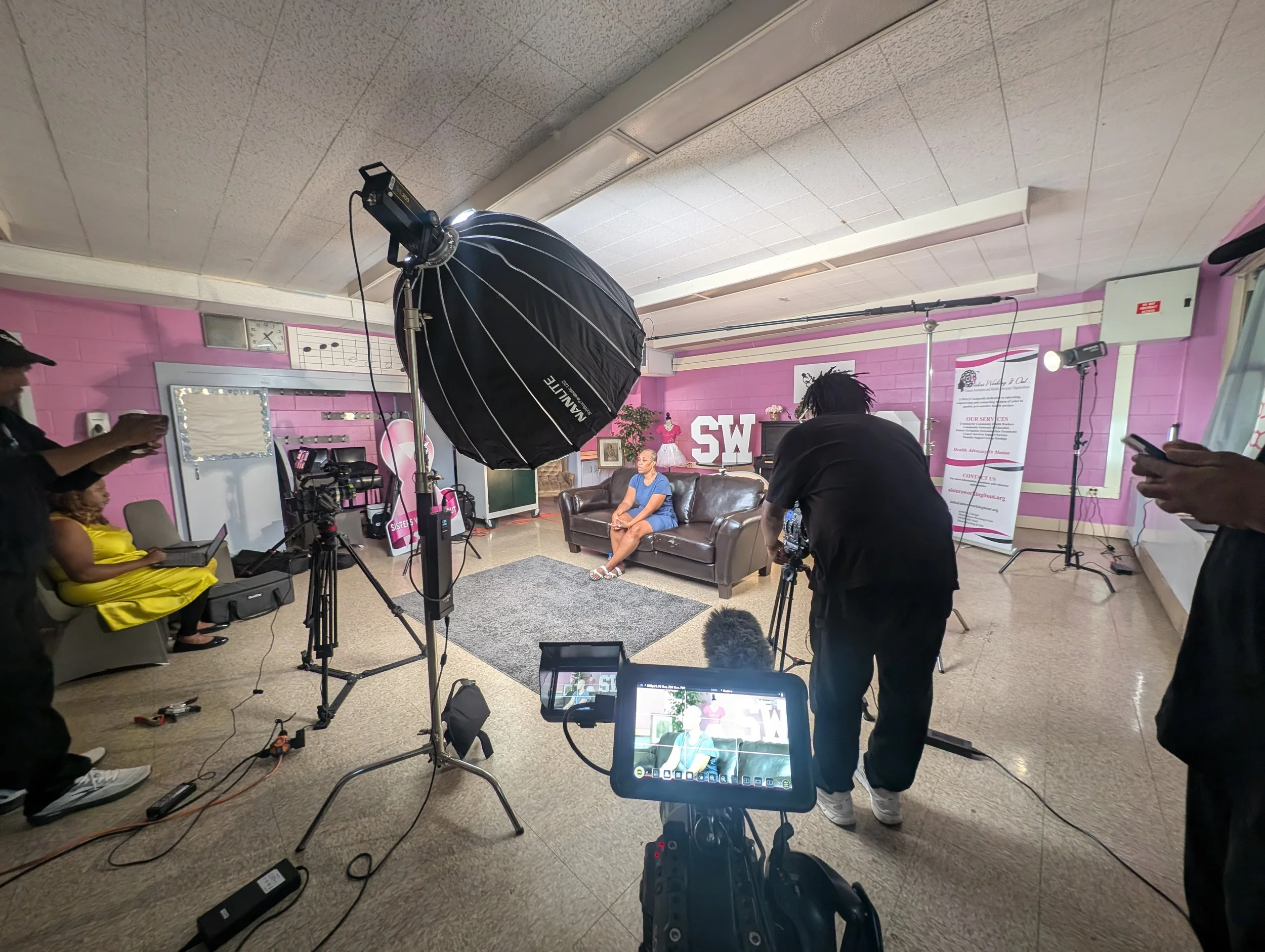 A woman in blue scrubs is seated on a brown leather sofa in front of a pink brick wall with large white letters reading 'SW'. She is being filmed by a camera crew and photographers in a room with pink walls and studio lighting. There are various piec