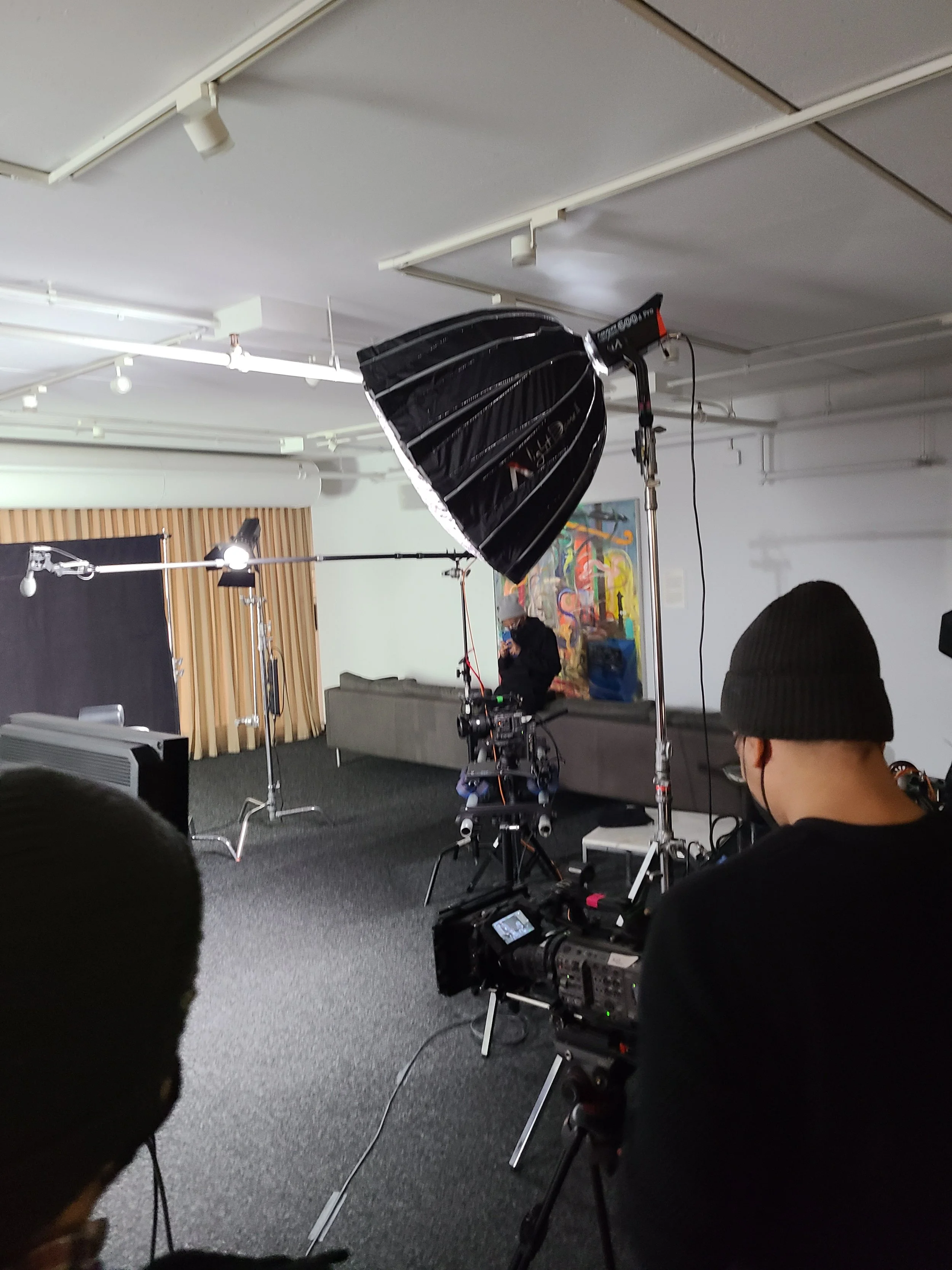 Filming studio with cameras, lighting, and a backdrop, featuring crew members preparing for a shoot in an art-filled room.