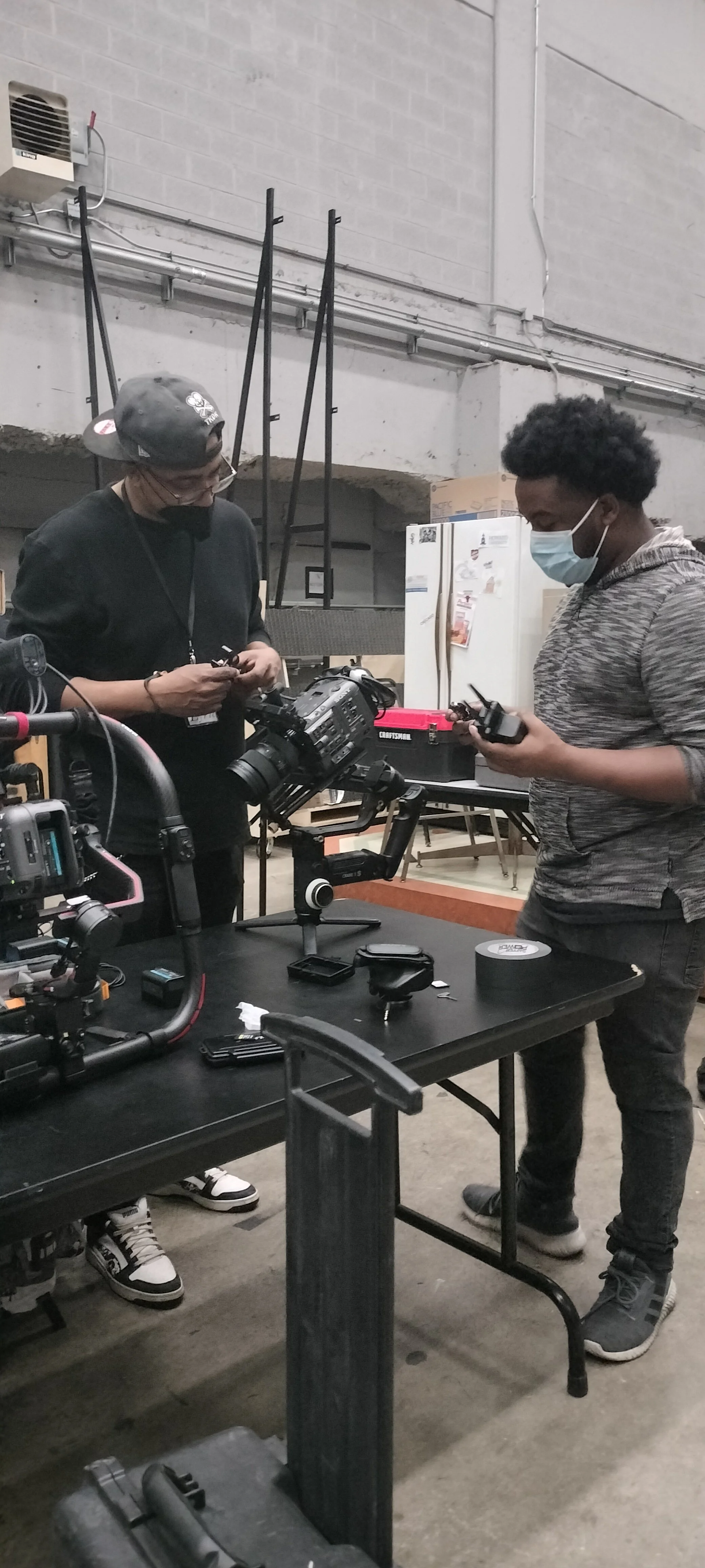 Two young men wearing face masks are working with camera equipment on a black table in an industrial-style room. One is wearing a black cap and black shirt, and the other has curly hair and a grey hoodie. There are various camera gear and tools on th
