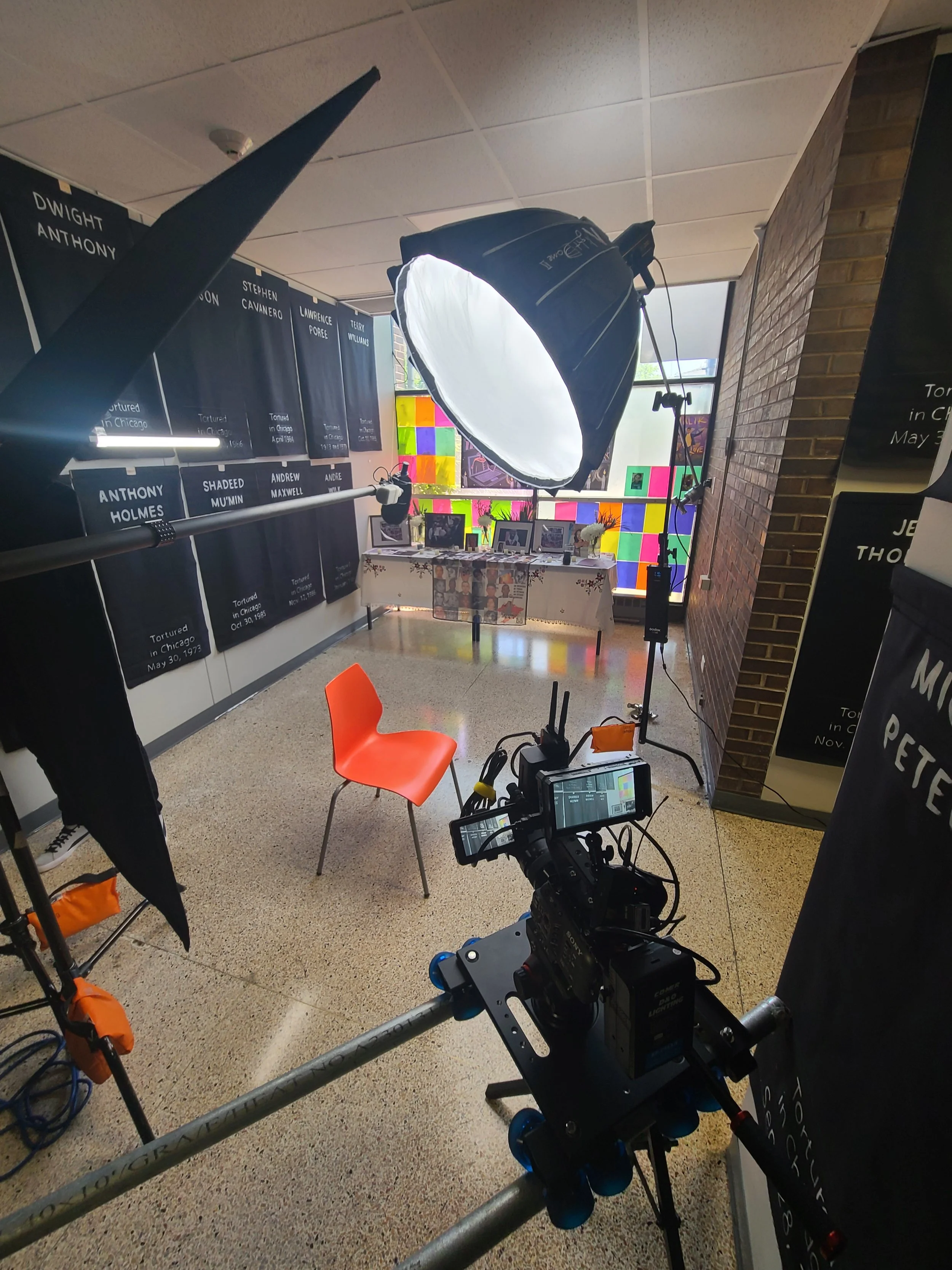Photography setup with a camera on a dolly, a large light with a softbox, an orange chair, and colorful stained glass windows. There are black banners with white text on the left wall and a decorated table with framed photos in the background.