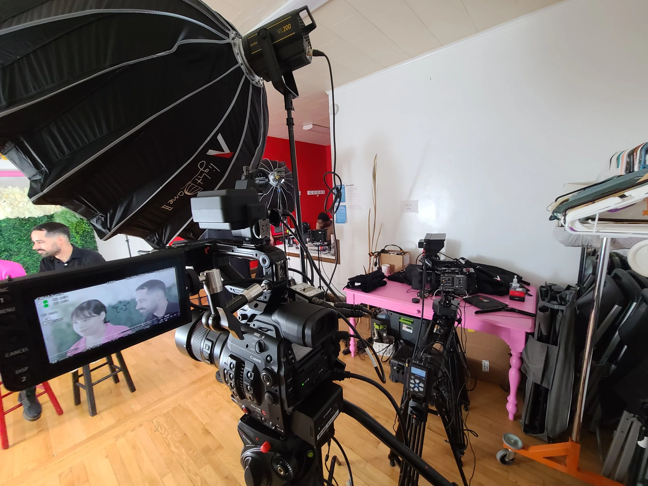 Video production set with camera, lighting, and crew preparing for a shoot featuring two people on a pink backdrop.