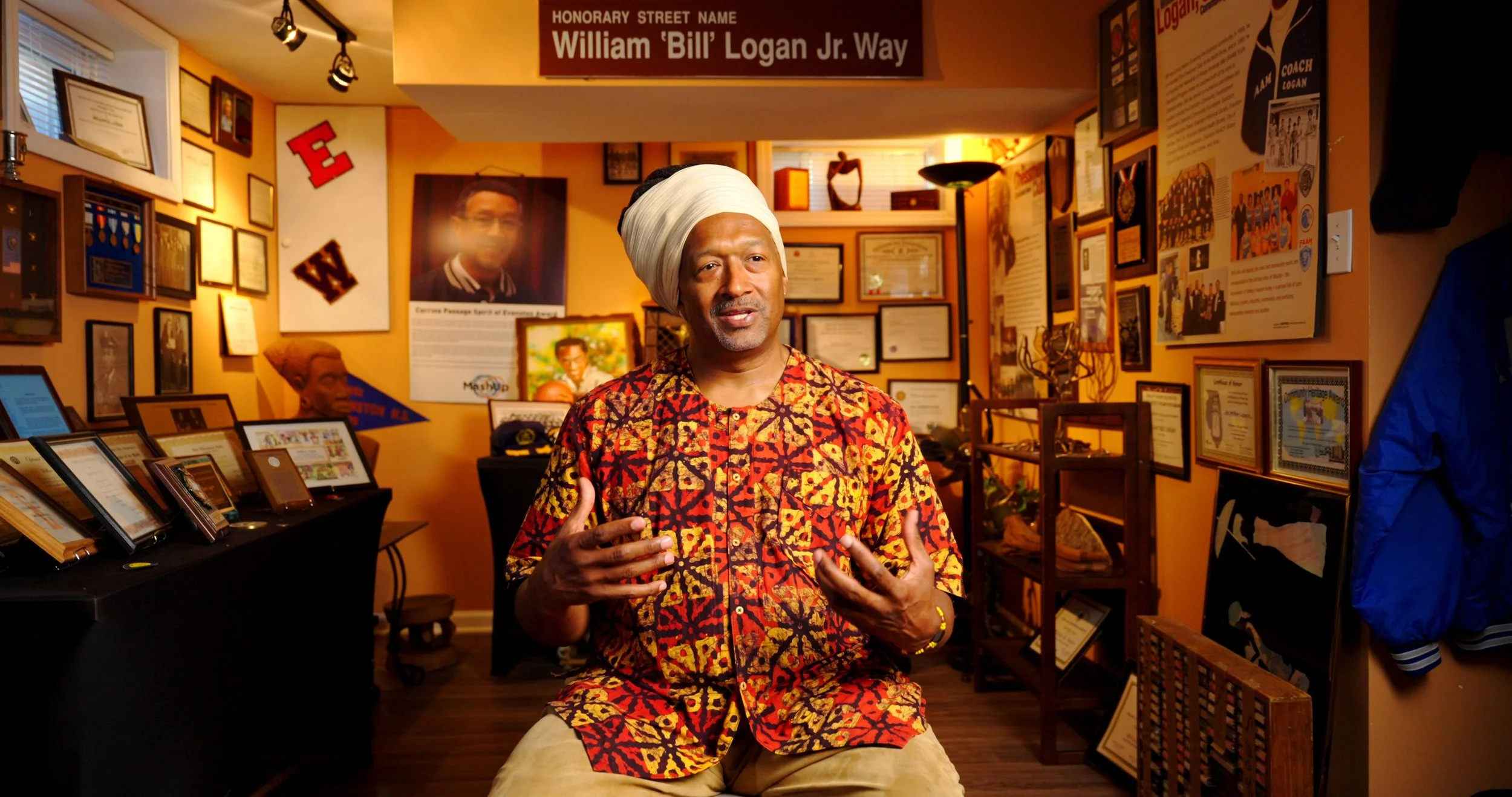 A man wearing a colorful African print shirt and white turban sits in a room filled with awards, photographs, and memorabilia, speaking and gesturing with his hands.