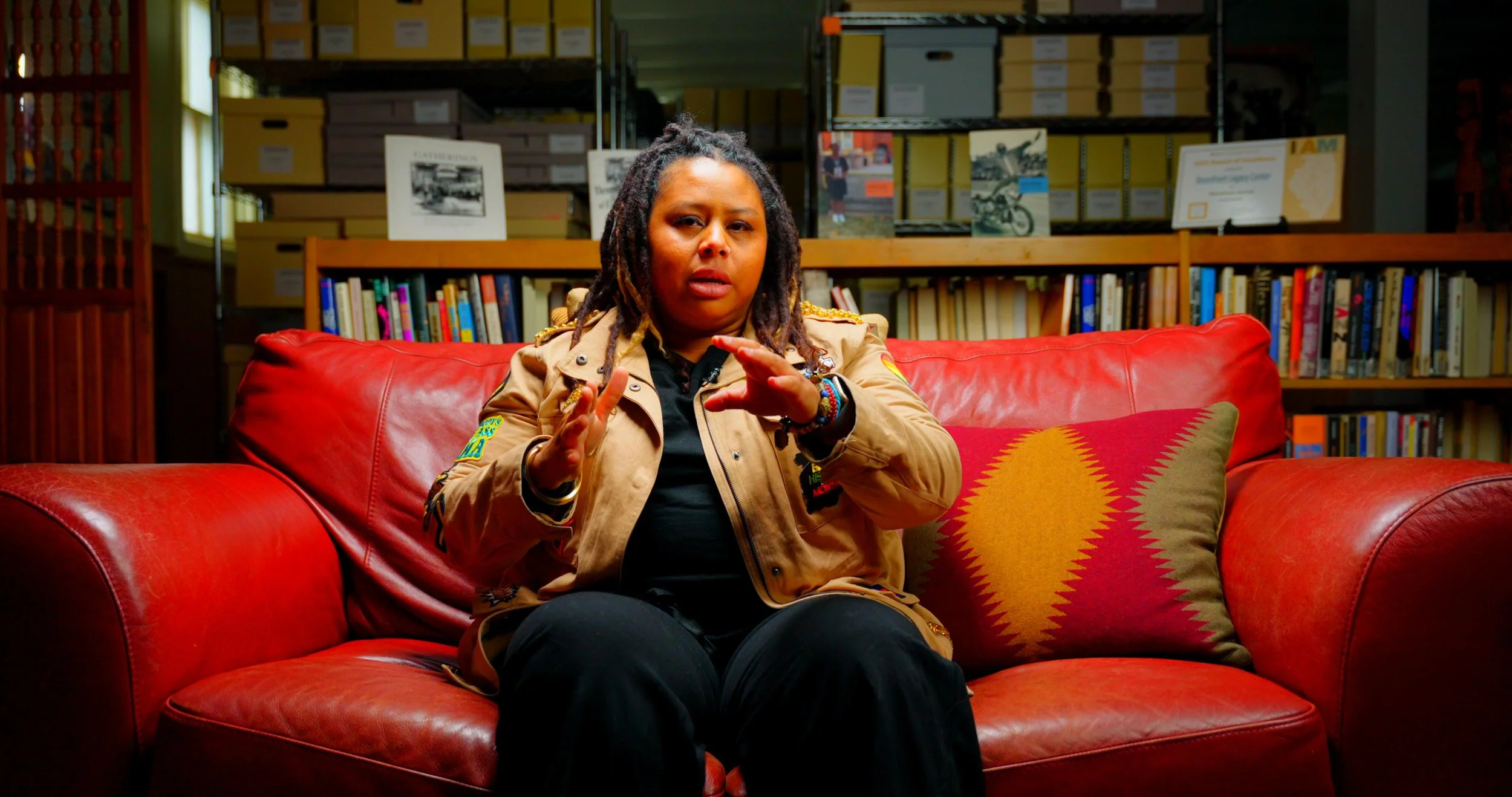 A woman with dreadlocks sitting on a red leather couch in a room with bookshelves in the background, gesturing with her hands.