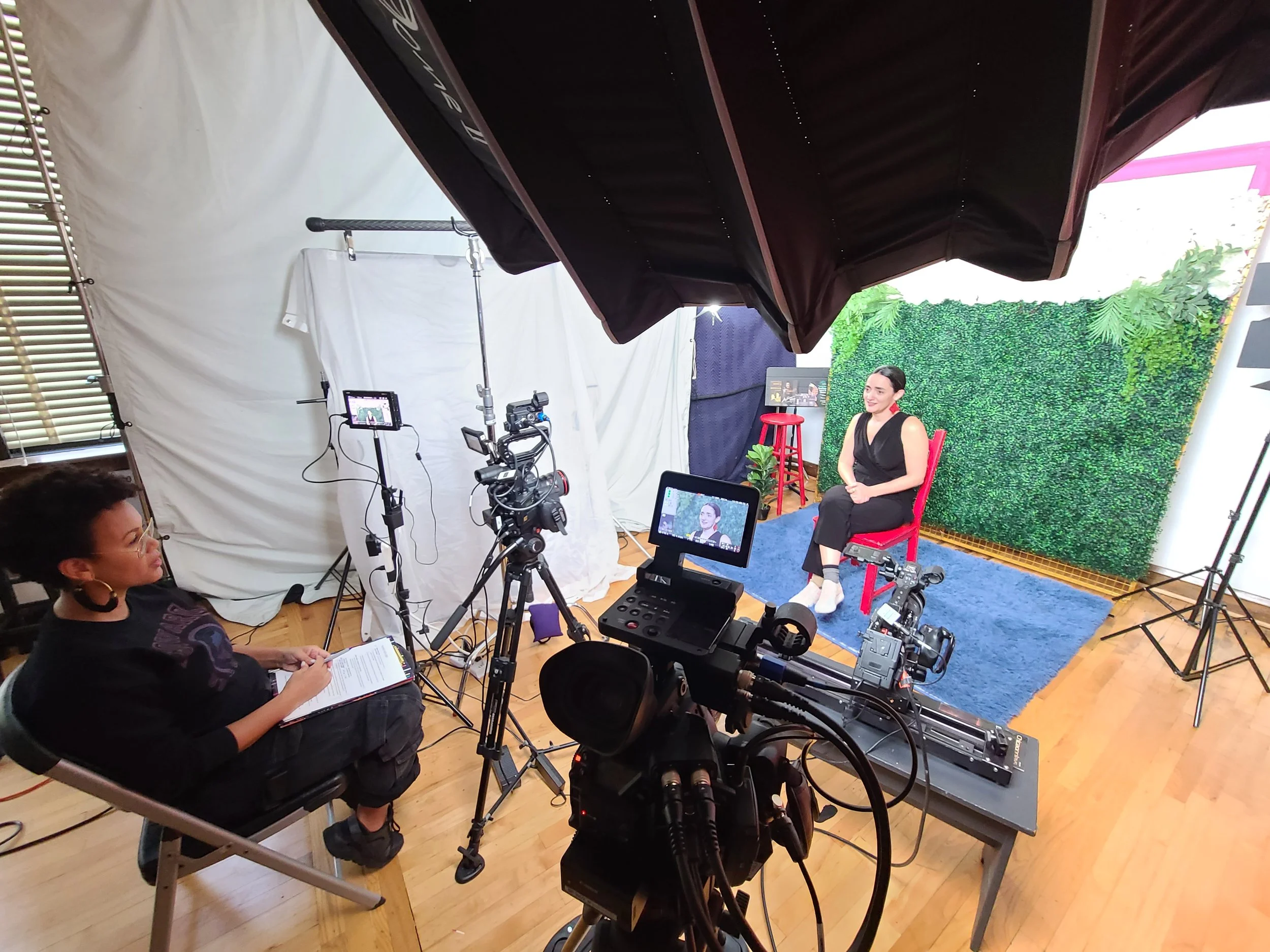 A woman in a black outfit and white shoes is seated on a red chair in a video recording studio, with a green leafy backdrop behind her. A director or producer is seated on the left, taking notes, with cameras, lights, and equipment set up around them
