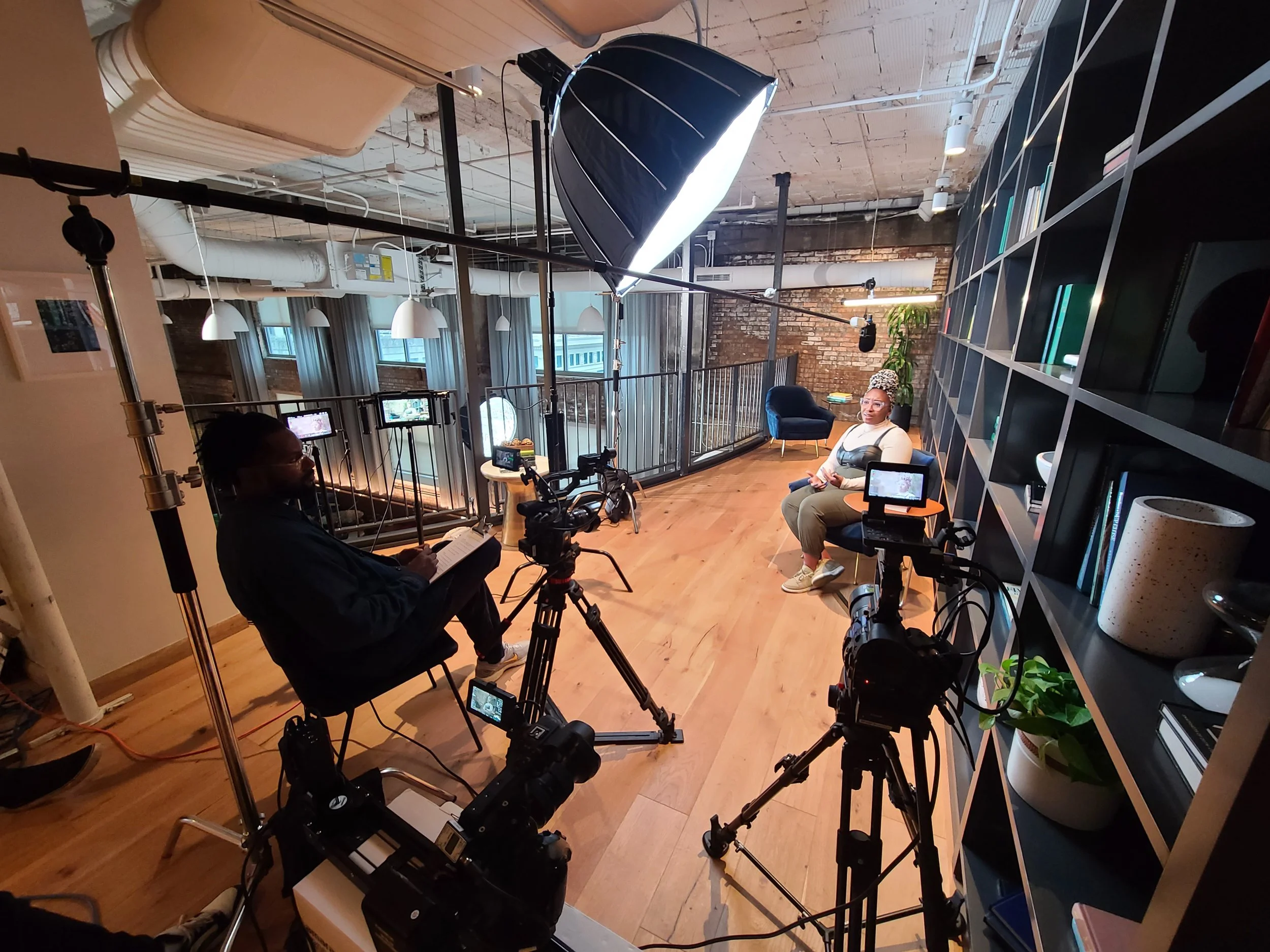 A woman sits in front of a camera in a recording studio with video production equipment, including cameras, lights, and a large bookshelf, in an industrial-style room with brick walls and large windows.