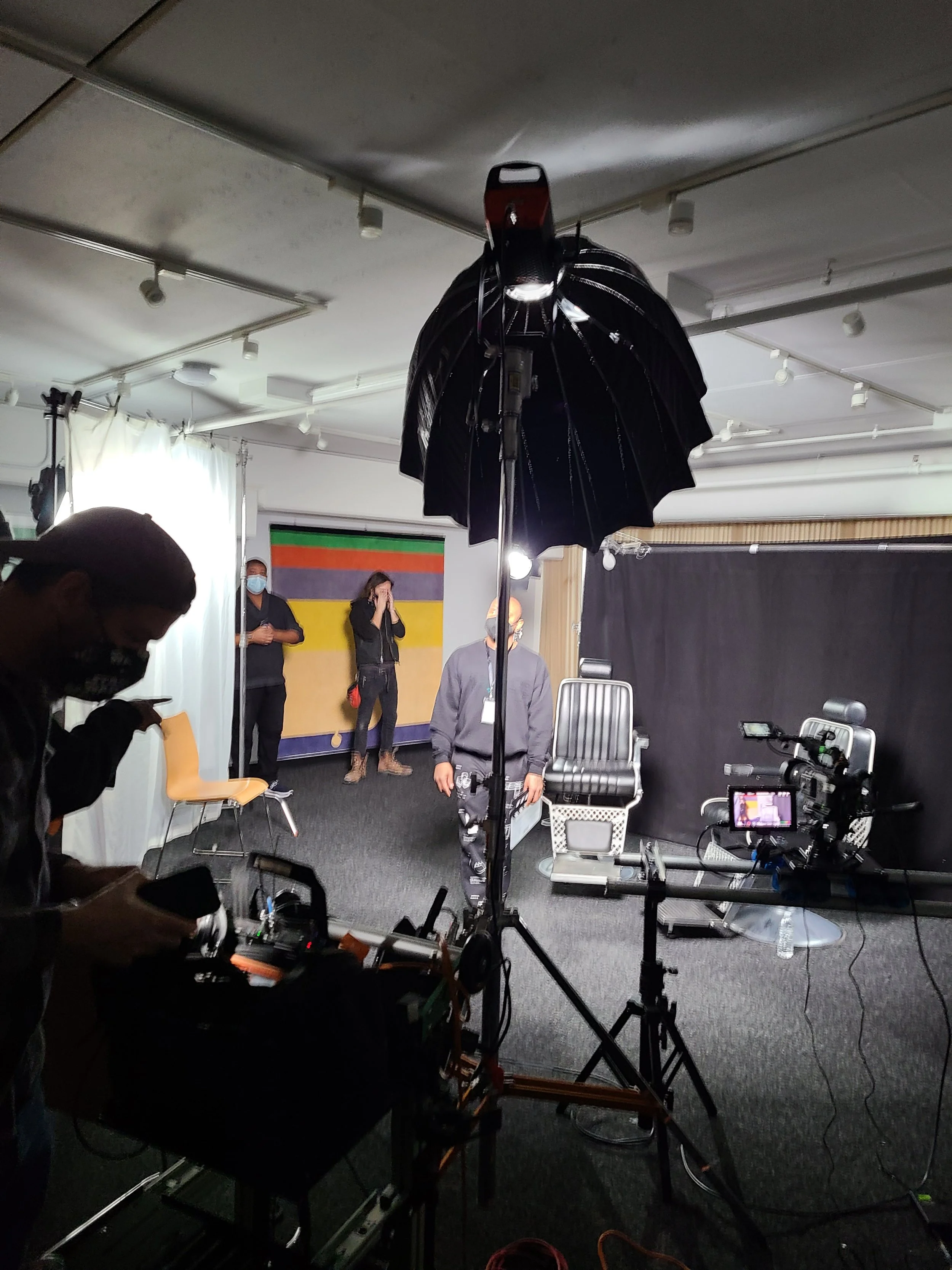 A video recording studio with a camera on a tripod pointed at a person standing in front of a backdrop with colorful stripes. There are lights, umbrellas for lighting, and crew members working around.