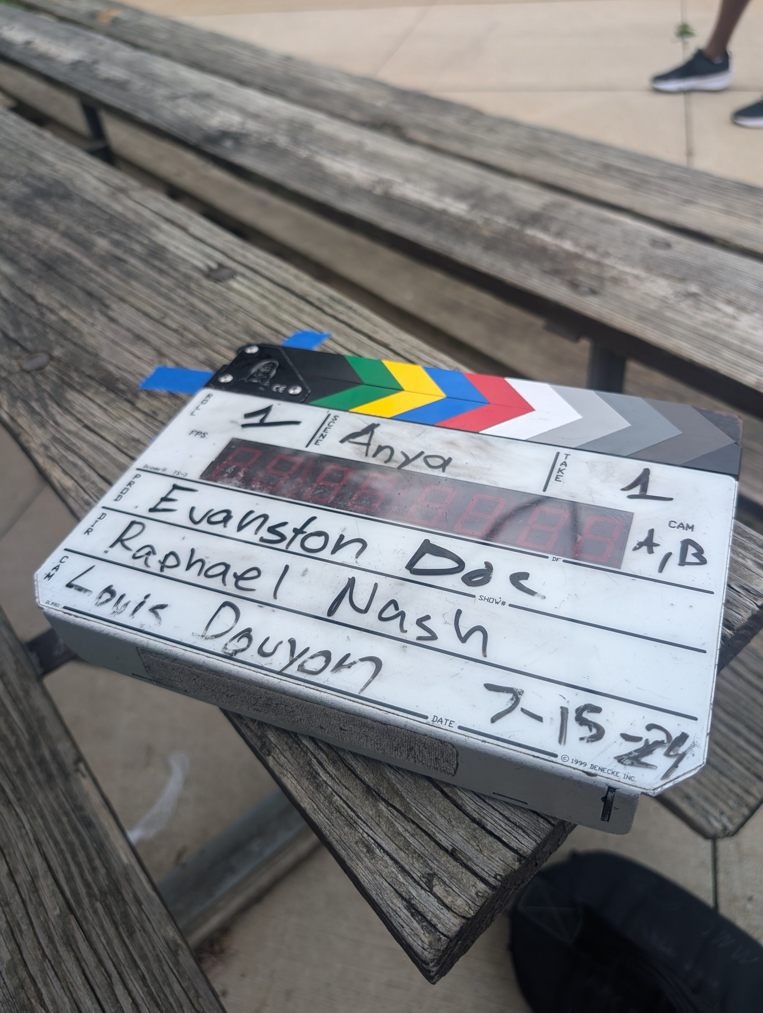 A film clapperboard on a weathered wooden table, with hand-written details including the names of cast members and the date, and a set of colorful items in the background.