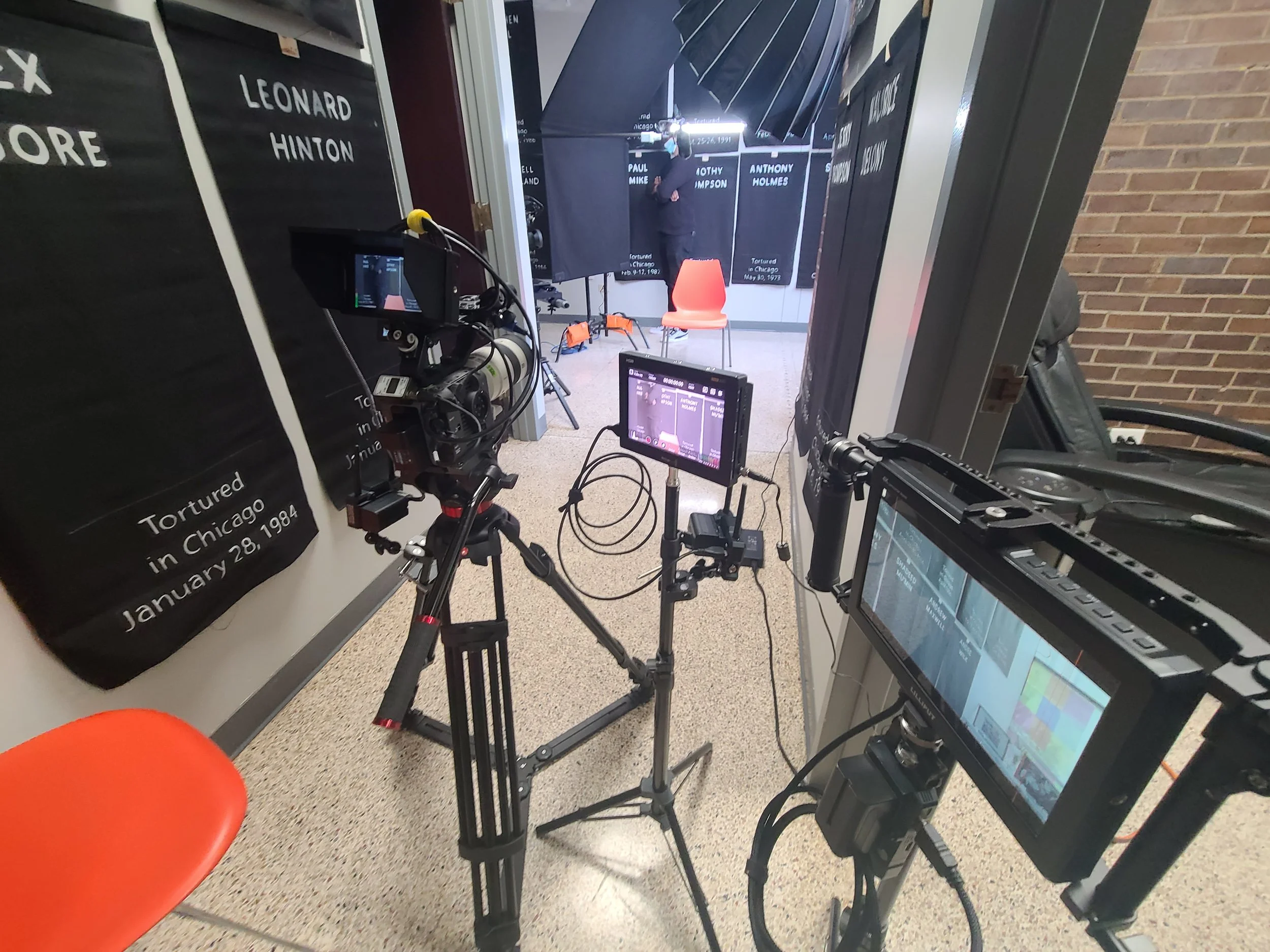 A recording studio setup with cameras and monitors aimed at a small stage with a red chair, backstage posters on the wall, and a man preparing for a recording or interview.