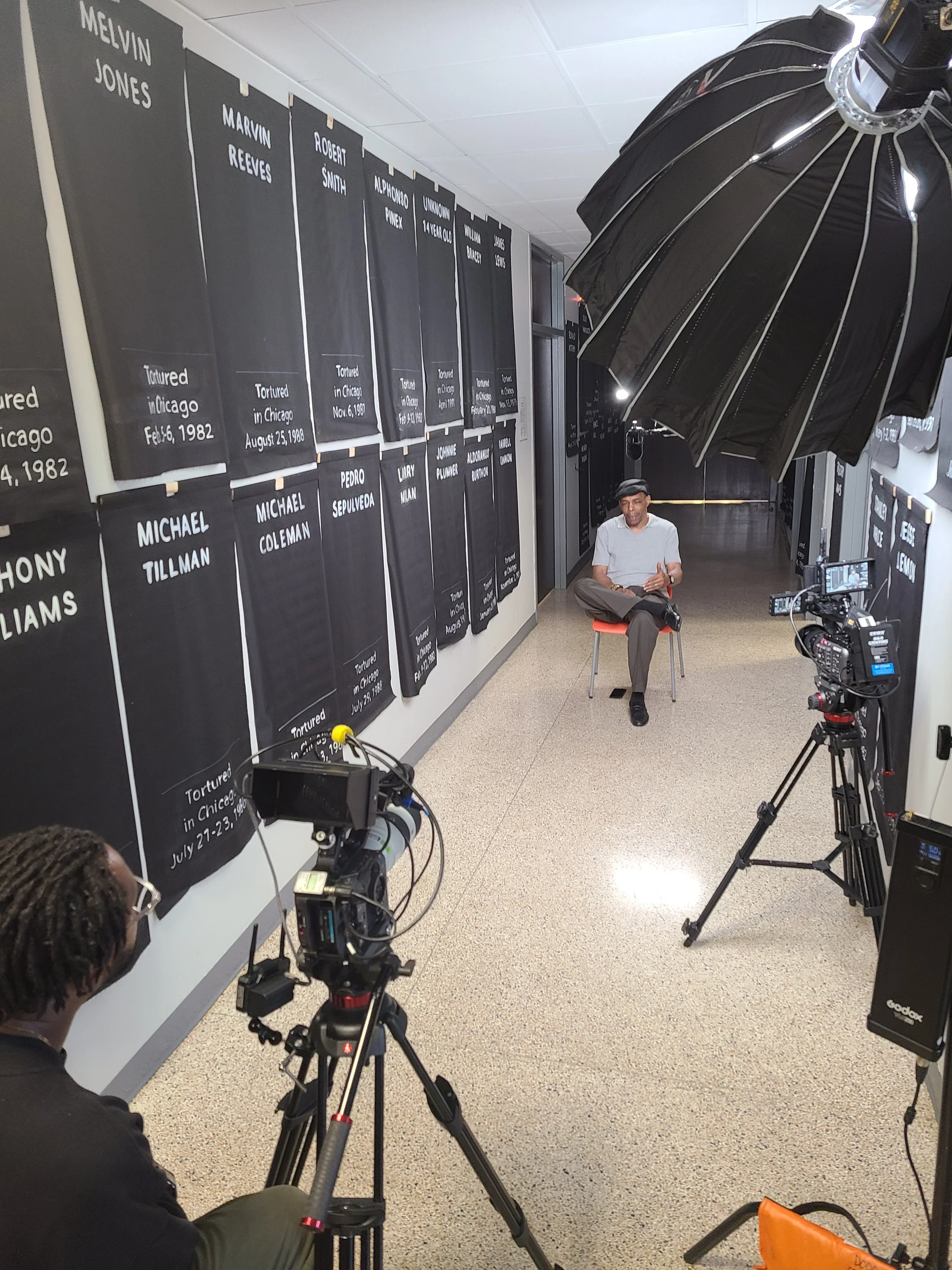 A man in a white shirt and gray cap seated in front of a wall of black memorial plaques with white text, being filmed or photographed with professional camera equipment, in a hallway with a black umbrella light above